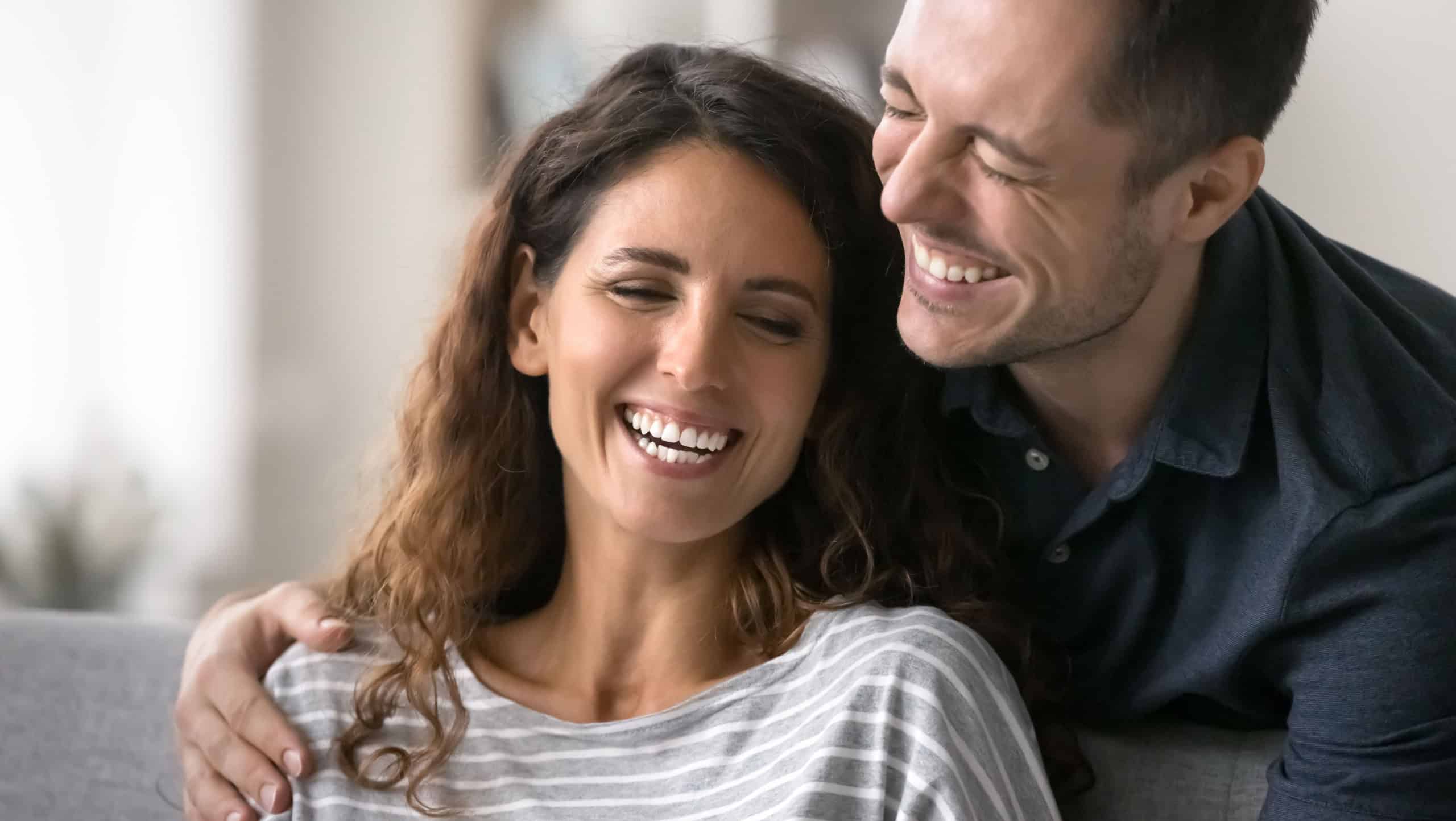 Close up beautiful overjoyed Latin couple talking, laughing, spend time together at home. Cheerful husband hugs his beloved beautiful wife, joking, giggling, feel carefree and happy. Relations, love