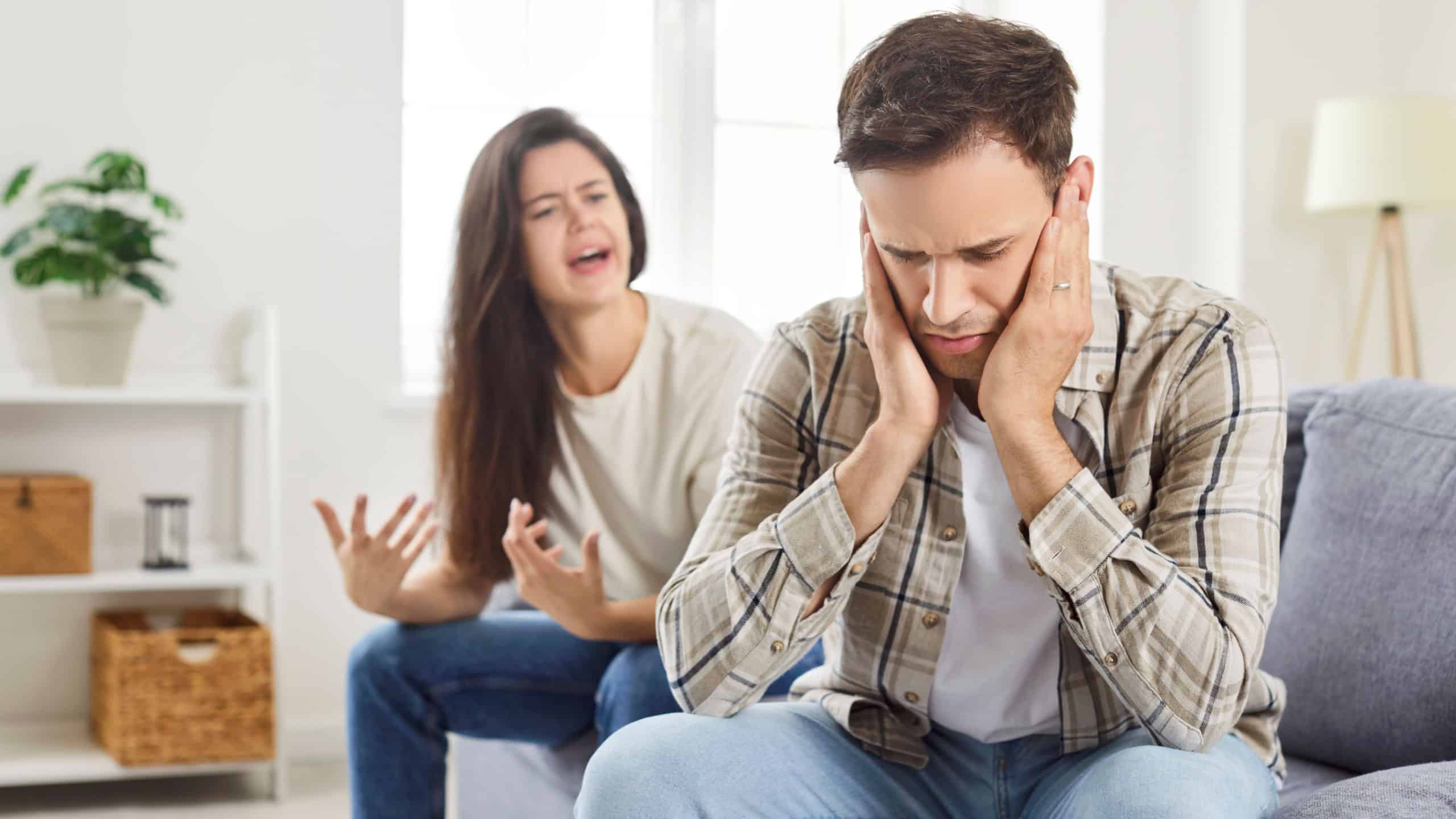 A young man sitting on the couch covers his ears from a womans scream. The wife scolds her husband, emotional problems in the family, the couple quarrels, relationship problems, disagreement