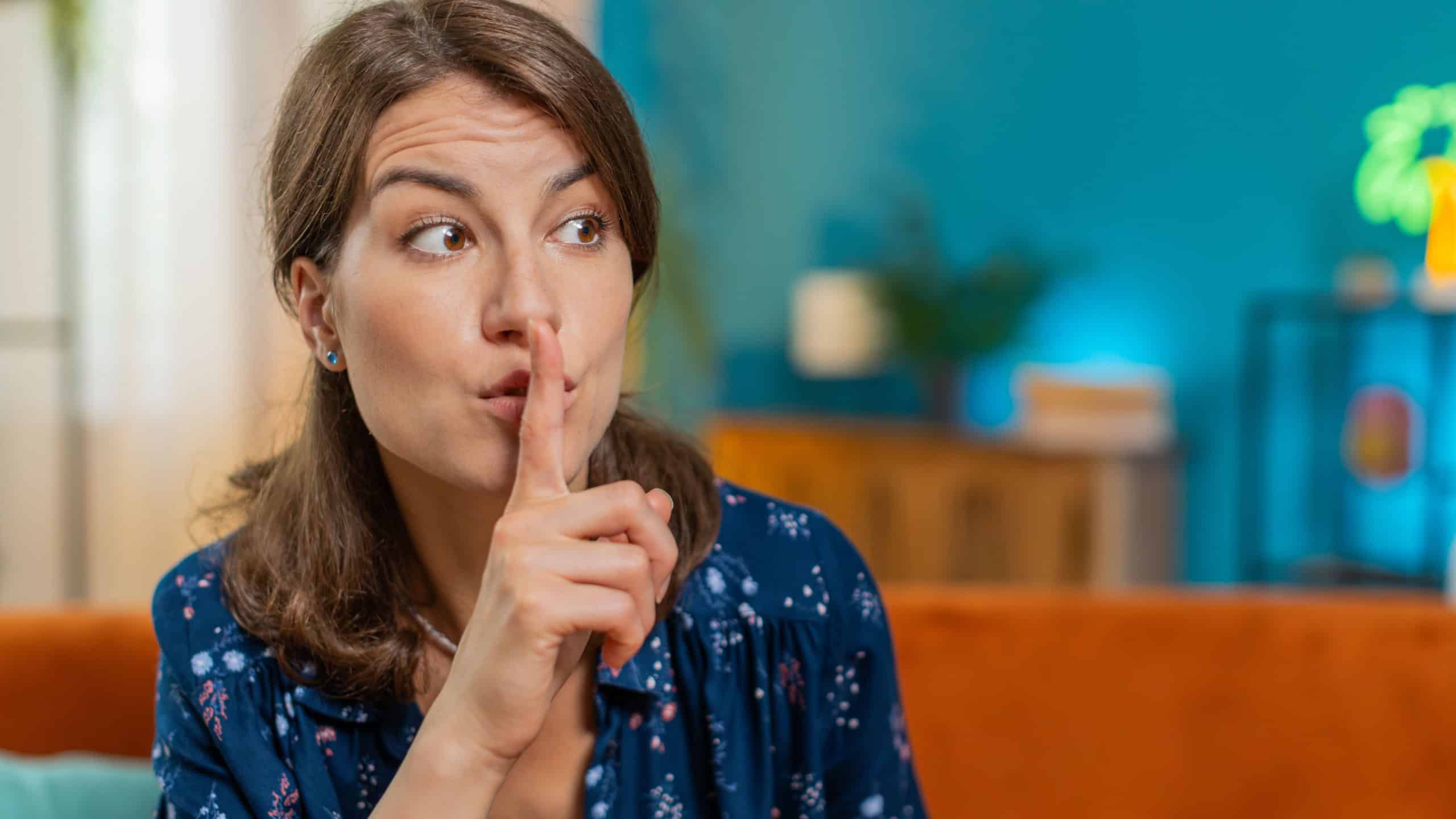 Shh be quiet please, silent. Portrait of young woman presses index finger to lips makes silence hush gesture sign do not tell gossip secret at home room apartment. Pretty girl sitting on couch sofa.
