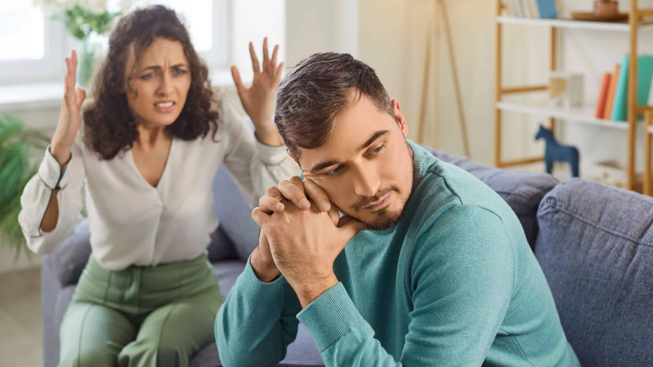 Guilty sad young man sitting on sofa, avoiding eye contact while angry upset woman shouting on him. Family couple arguing at home, having relationship problems, expressing anger and disappointment.