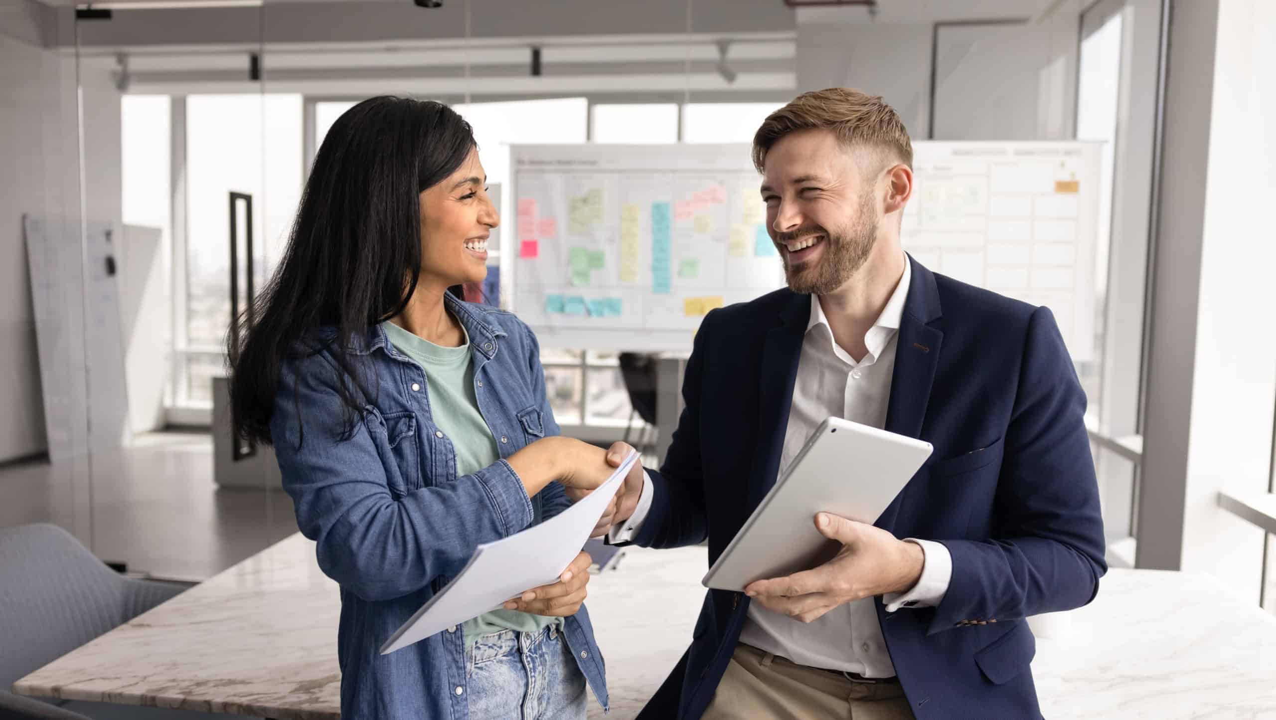 Positive multiethnic couple of coworkers shaking hands in office space, holding tablet, documents, smiling, laughing. Caucasian businessman thanking female Indian coworker for professional help