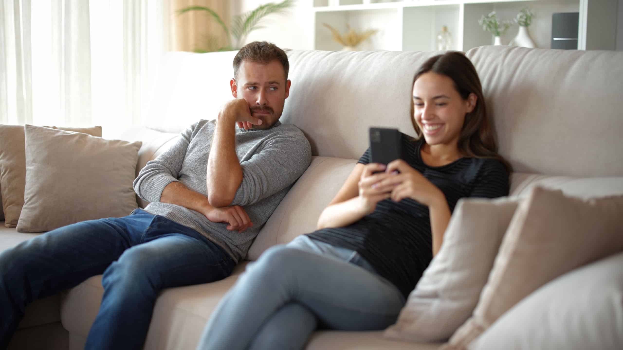 Worried man being ignored by his partner who is chatting online on a couch at home