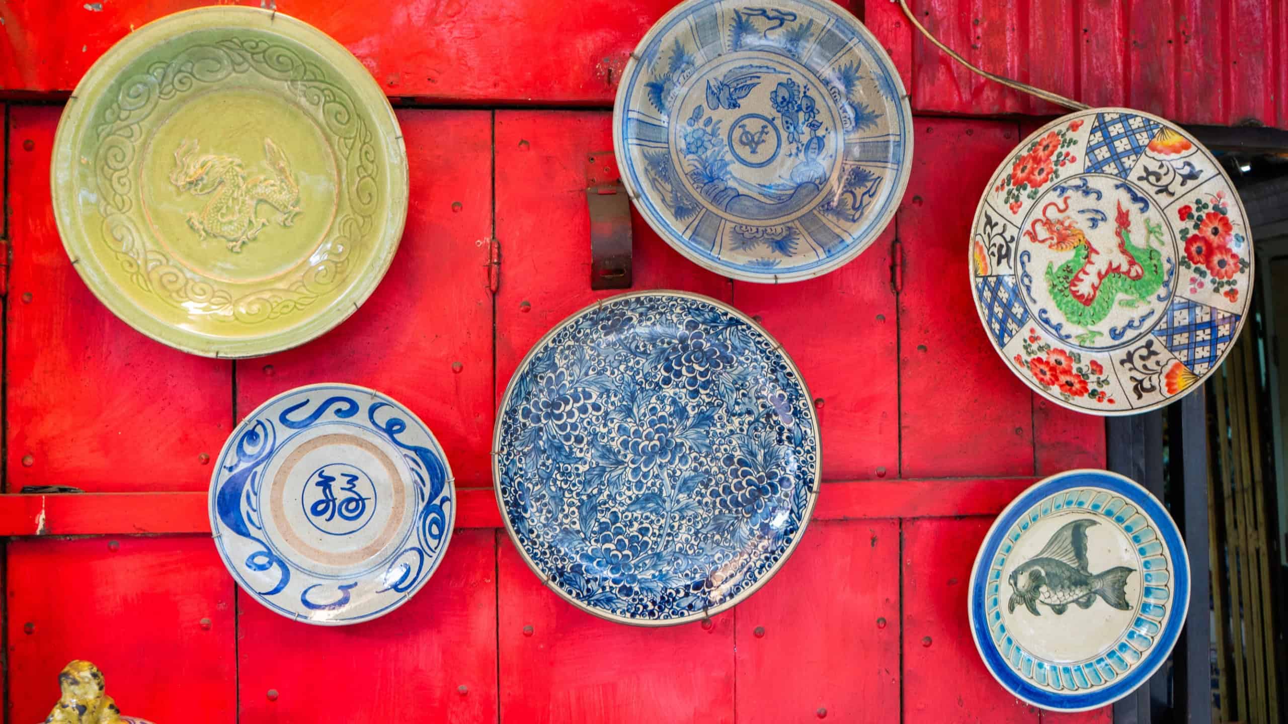 A collection of antique Asian ceramic and porcelain plates displayed as a decorative arrangement on a bright red wooden wall. Each plate features a unique traditional design