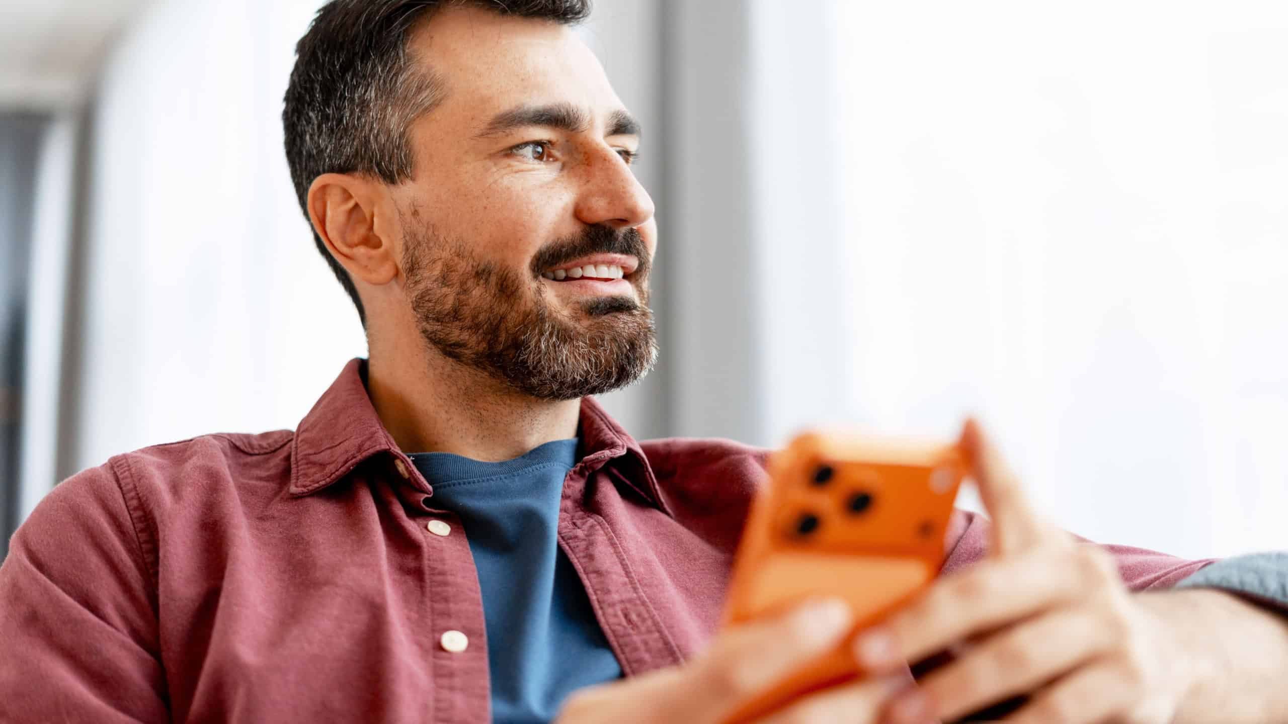 Smiling bearded man smiling and looking away while holding a modern smartphone, conveying casual digital connection, social browsing and relaxed confident lifestyle in studio setting