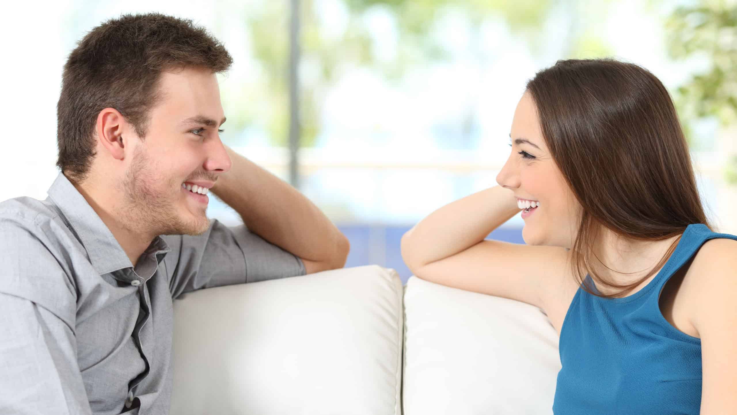 Side view of a couple talking and looking each other affectionate sitting on a couch at home with a window in the background