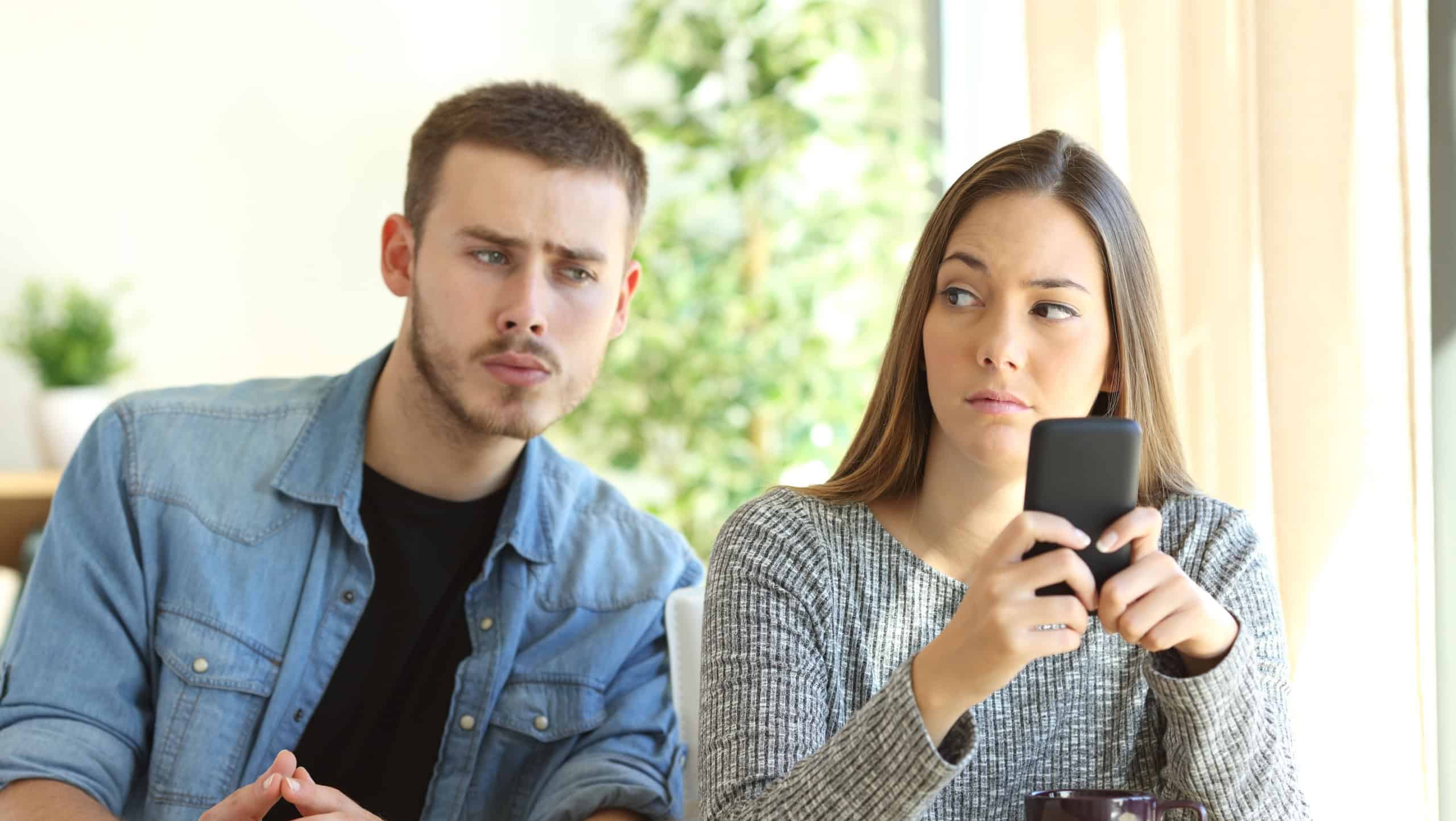 Jealous boyfriend spying his girlfriend watching her phone while she is looking him upset