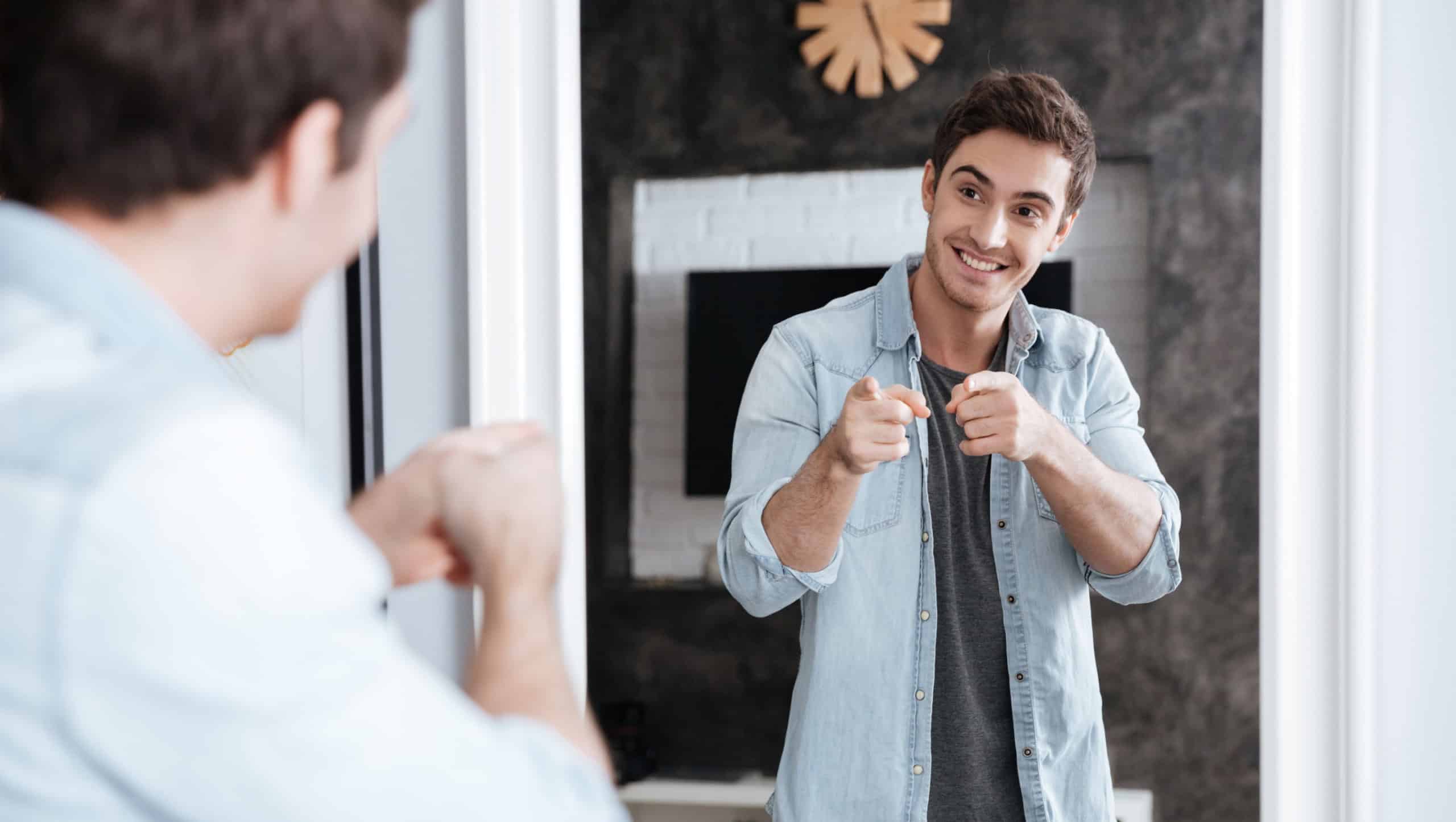 Smiling young man pointing fingers at his mirror reflection while standing at home