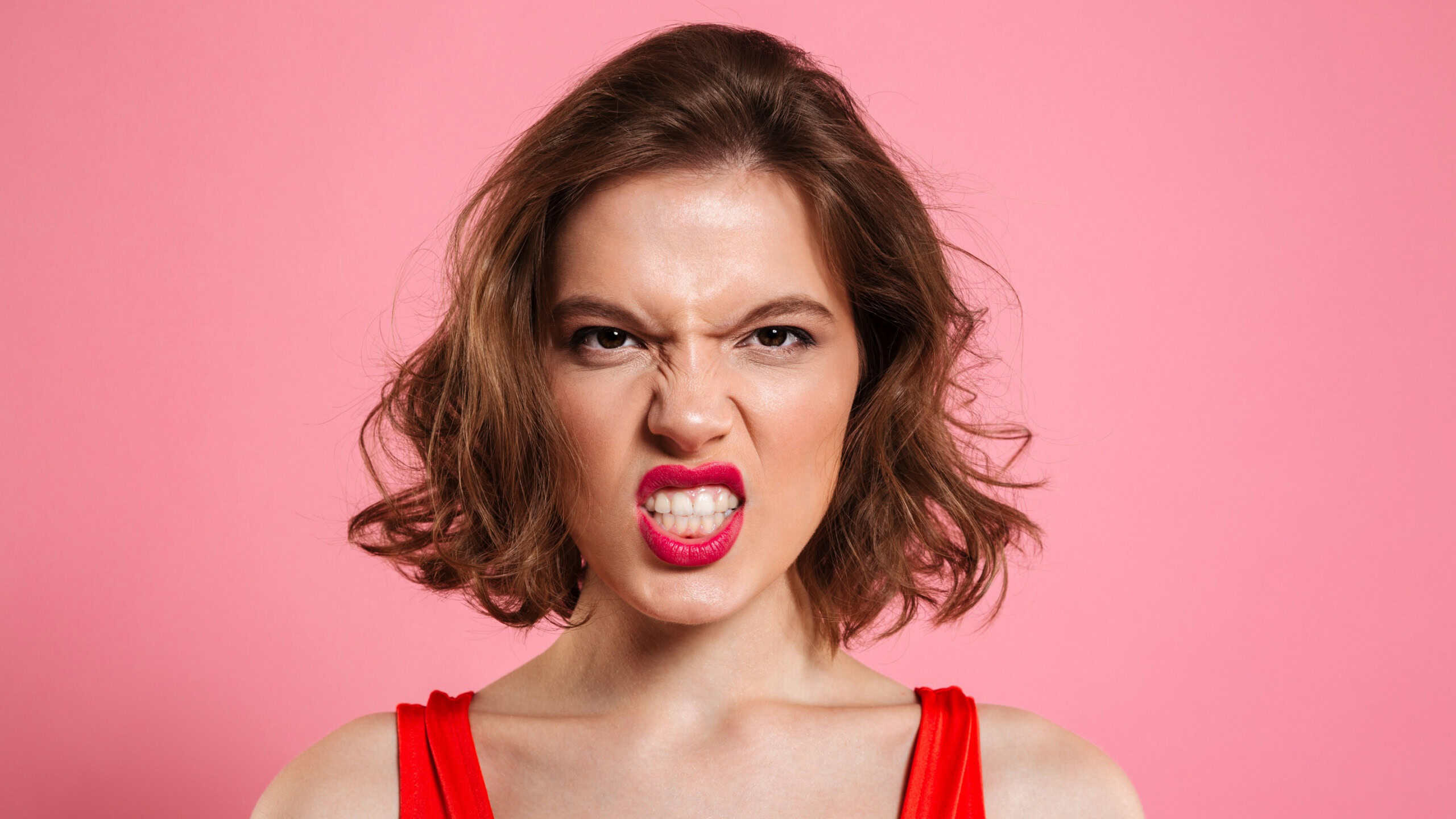Close-up portrait of angry young woman with red lips looking at camera, isolated on pink background