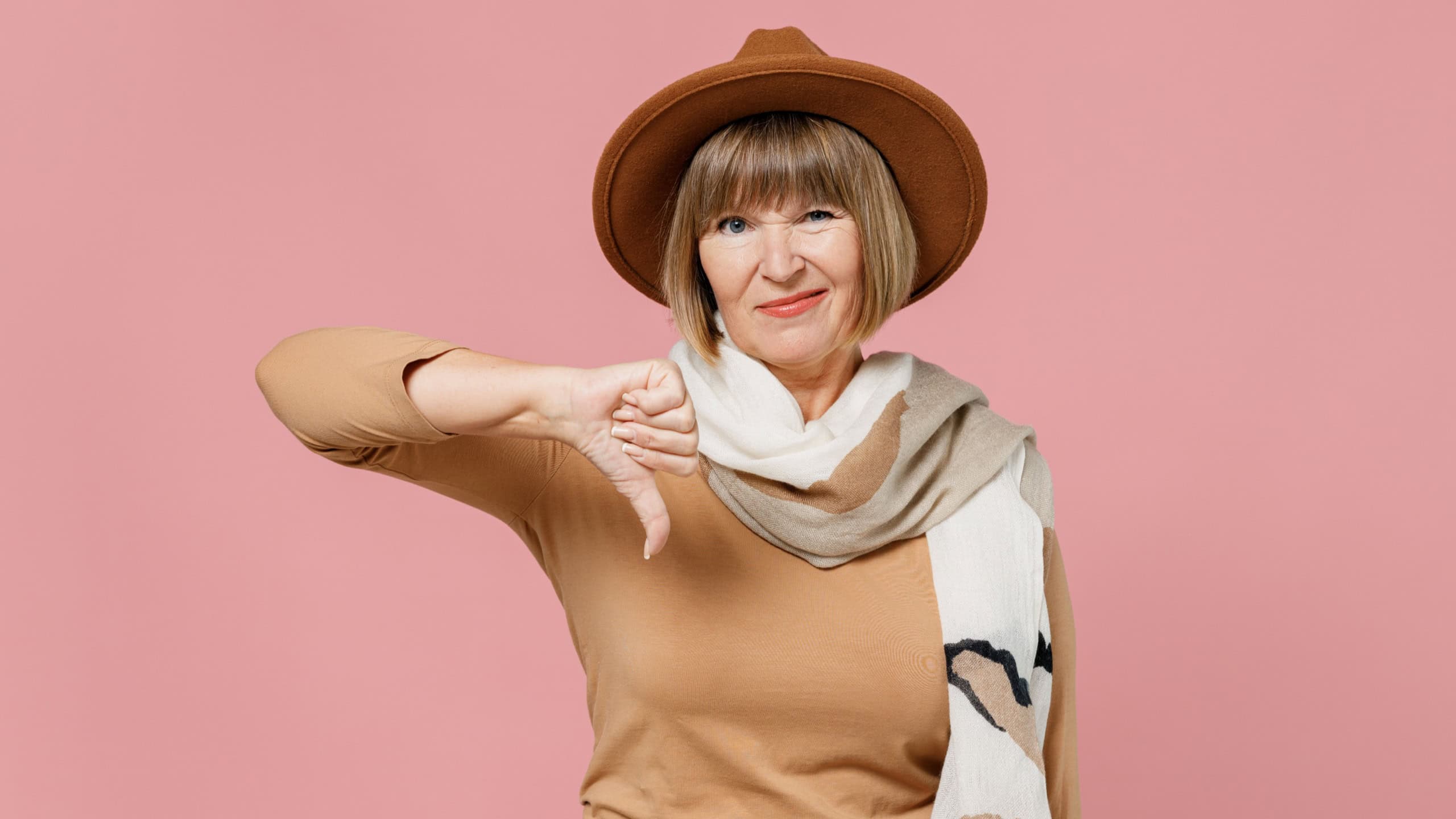 Traveler tourist displeased mature elderly senior lady woman 55 years old wears brown shirt hat scarf showing thumb down dislike gesture isolated on plain pastel light pink background studio portrait