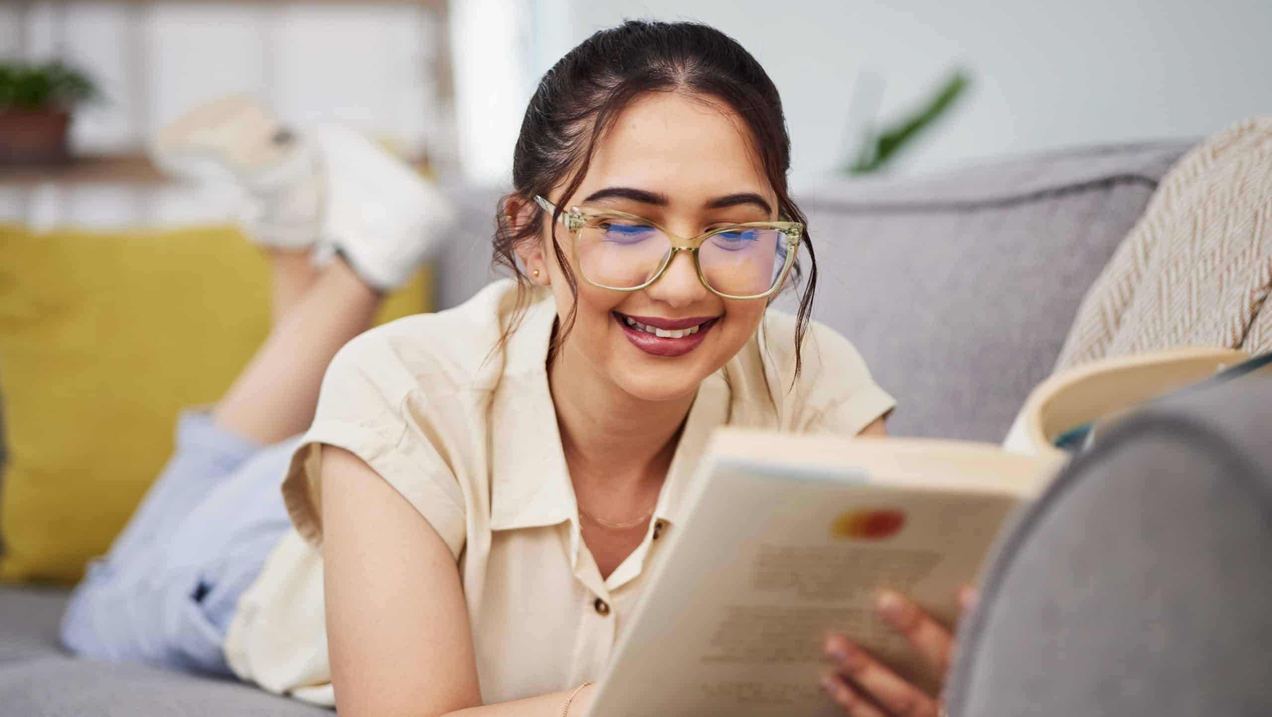 Happy woman, reading and book on sofa in home for story, novel and studying literature. Young student, gen z girl and relax with books in lounge for learning, knowledge and education in living room