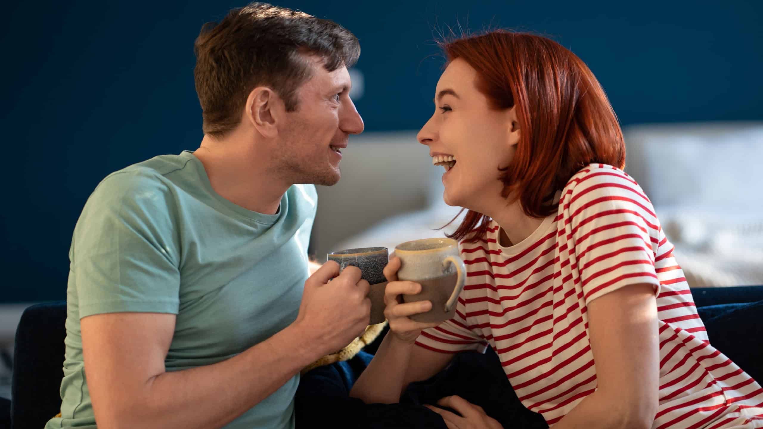 Joyful married couple laugh together morning with cup coffee sitting on couch. Smiling wife with husband enjoy holiday at home. Overjoyed European family have fun weekend tell each other news, jokes.
