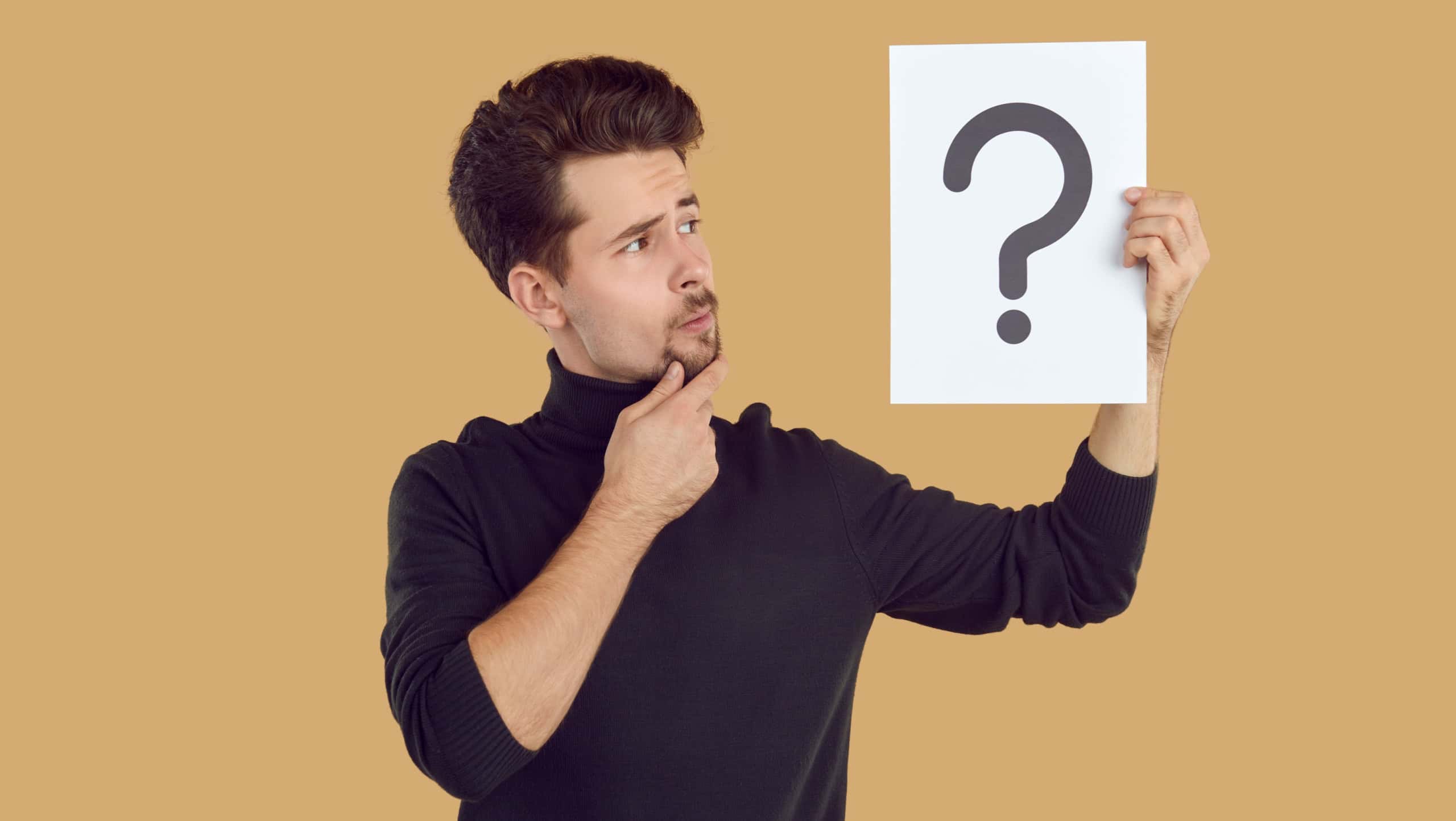 Pensive man is looking at piece of paper with question mark drawn on it, which he is holding in his hand. Young Caucasian man holding question mark symbol and thinking isolated on beige background.