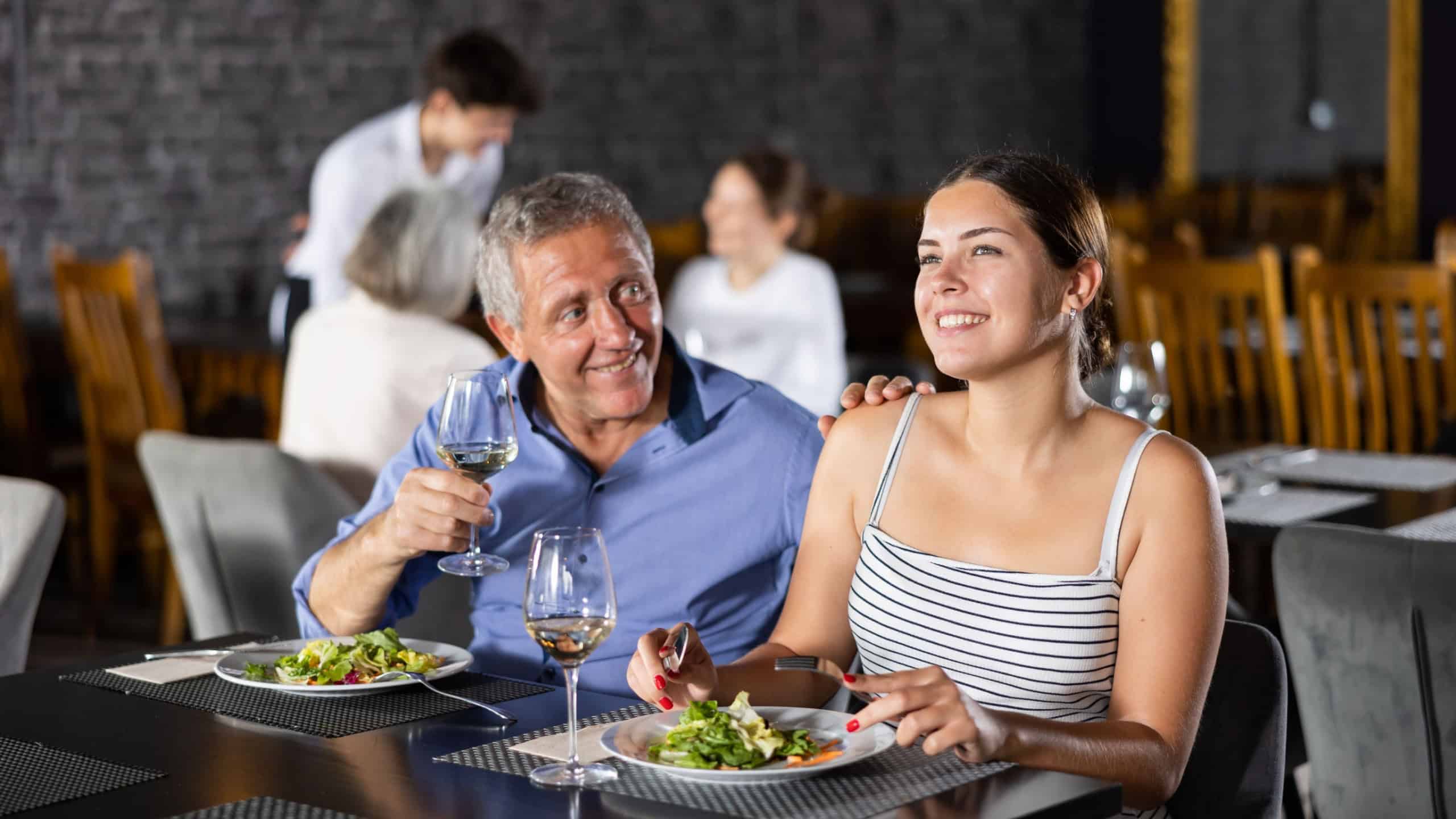 Smiling senior man spending time with young girl in restaurant. Carefree couple laughing merrily, enjoying delicious food and drinking wine at dinner. Age-gap relationships concept