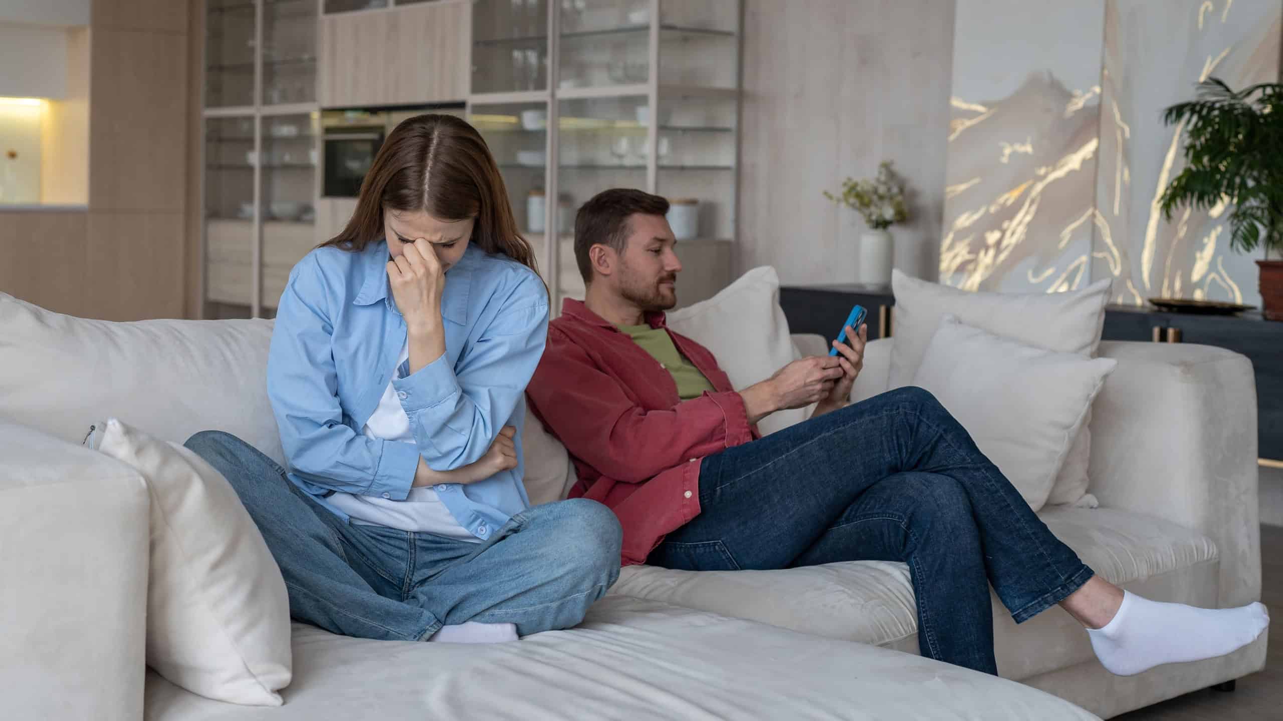 Indifferent man ignores crying woman, covering face with hand, cannot holding back tears. Sad wife sobs, turning back on husband. Uncaring boyfriend uses phone without paying attention to girlfriend