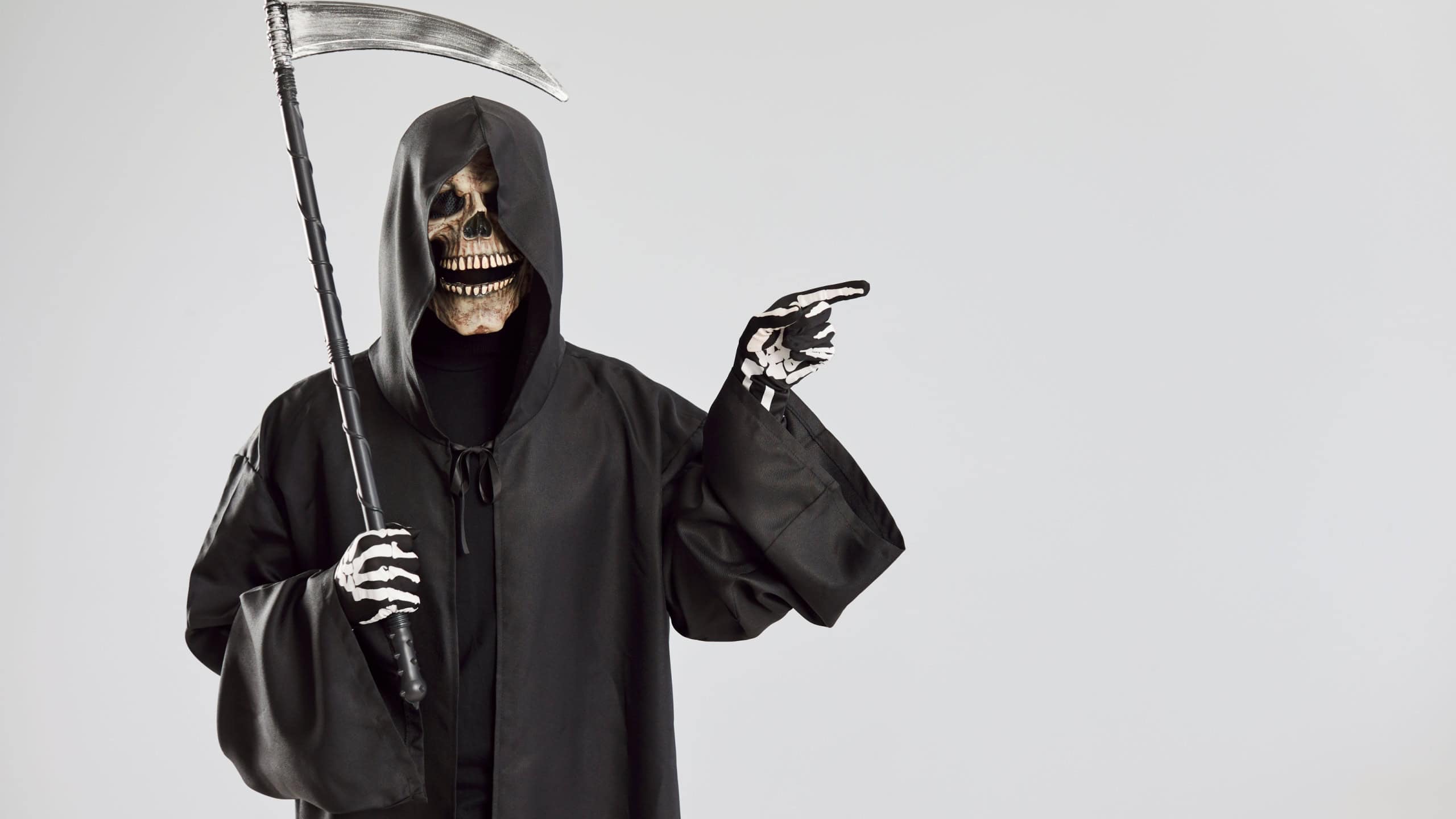 Male person in Halloween grim reaper death costume holding scythe in black cloak with hood pointing index finger to the side isolated on studio gray background. Halloween party and horror concept.