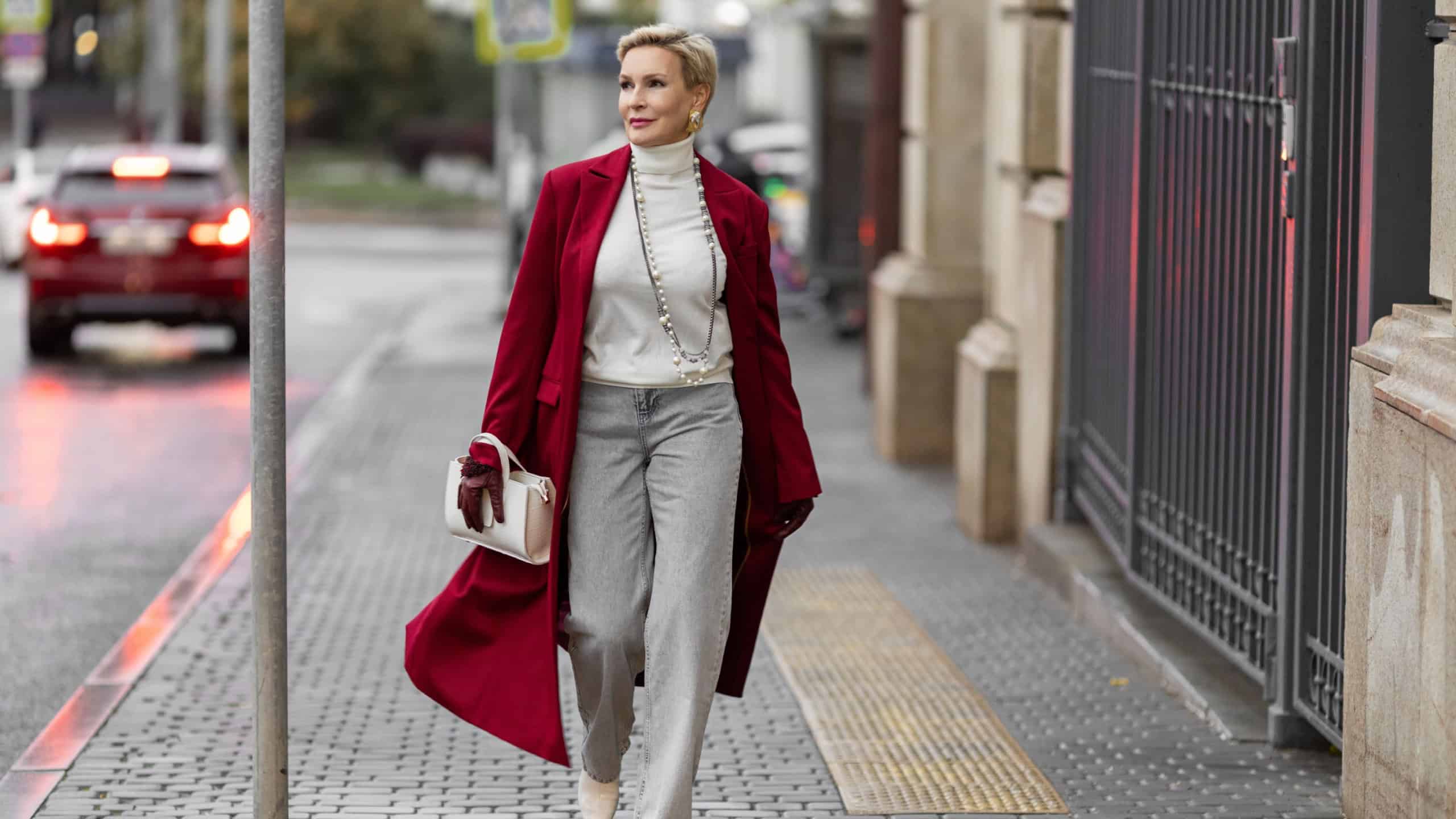 Stylish Woman in Burgundy Coat Walks City Street. Wearing Fashionable clothing White Turtleneck Sweater, Pearl Necklace and Grey Jeans, Carrying White Bag. Confident and Chic Mature Fashion Model