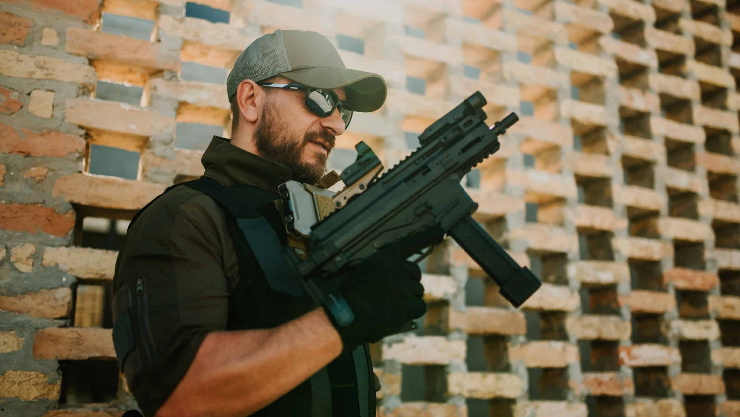 Private military contractor wearing tactical gear and sunglasses holds a submachine gun while standing near a brick wall in a conflict zone