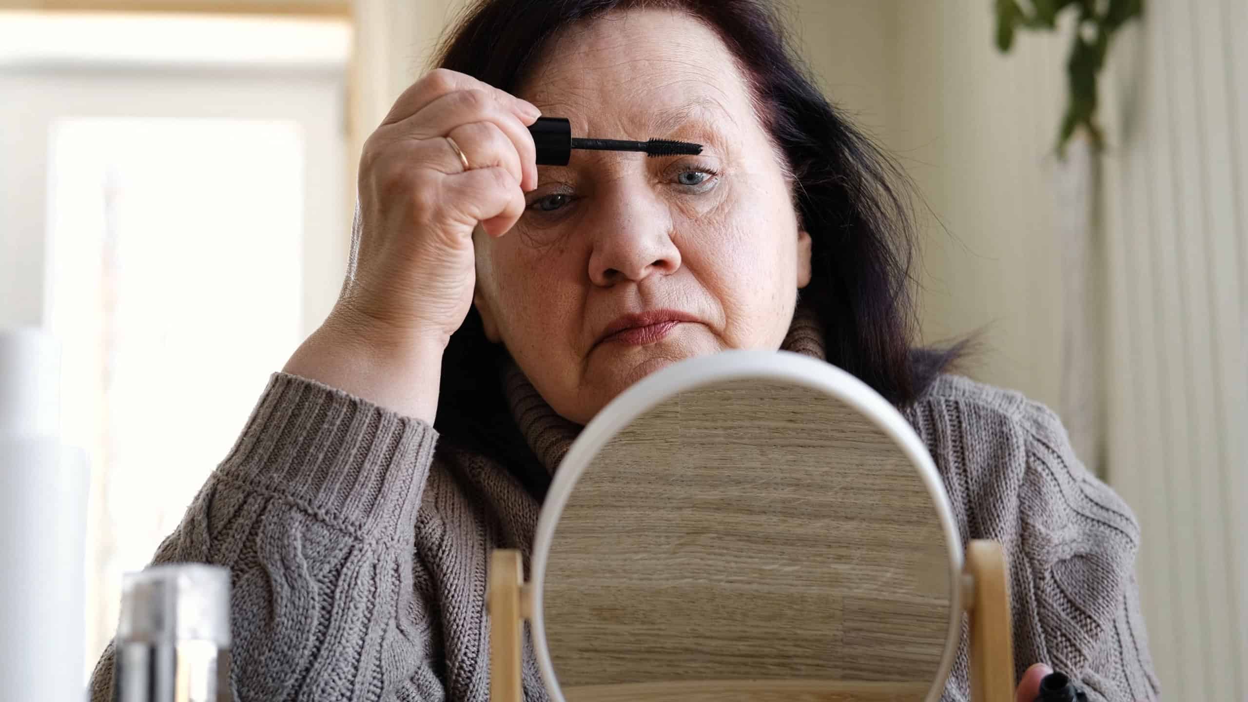 Attractive brunette senior woman putting on makeup looking in the mirror. Beautiful lady caring her face. Elderly female applied cosmetics. Natural old beauty and aging concept. Skin care and make-up