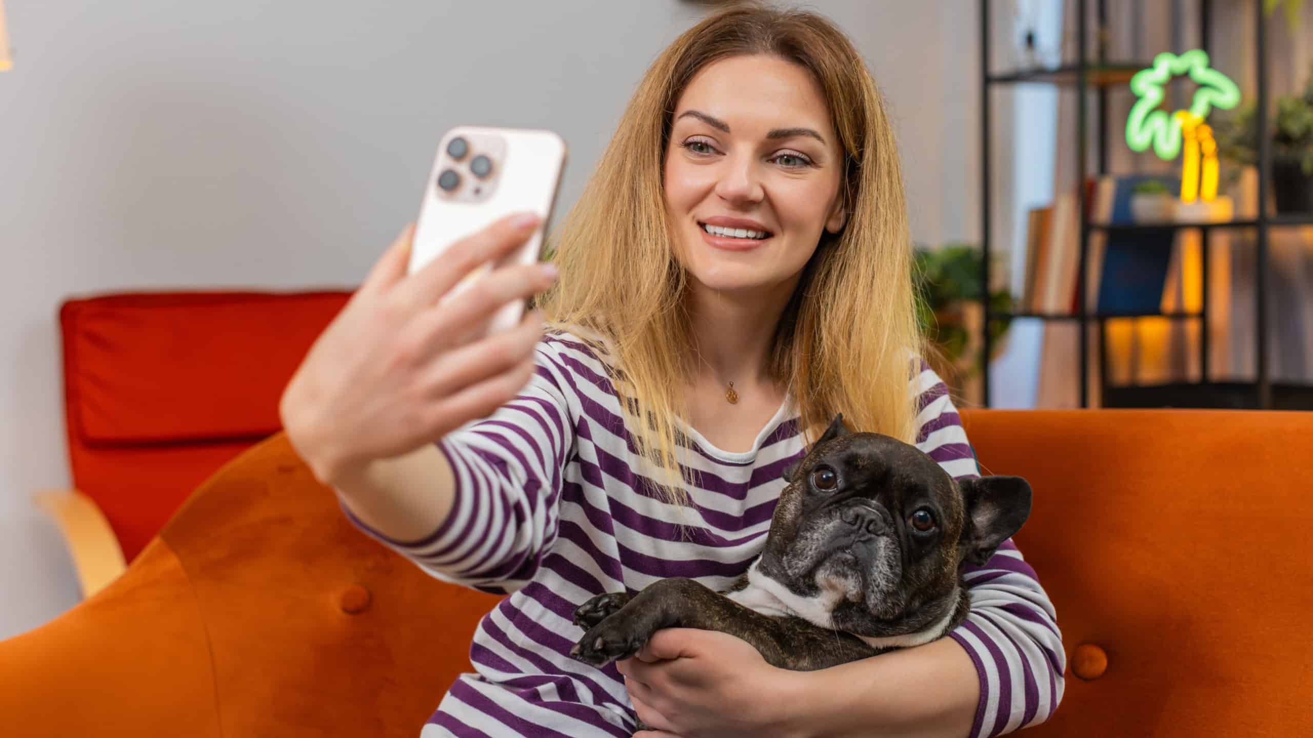 Middle-aged woman with French bulldog at home takes funny selfies on smartphone laughs happy mood playful style. Girl with dog on sofa joins video call makes faces and smiles shares joy chat fun live.