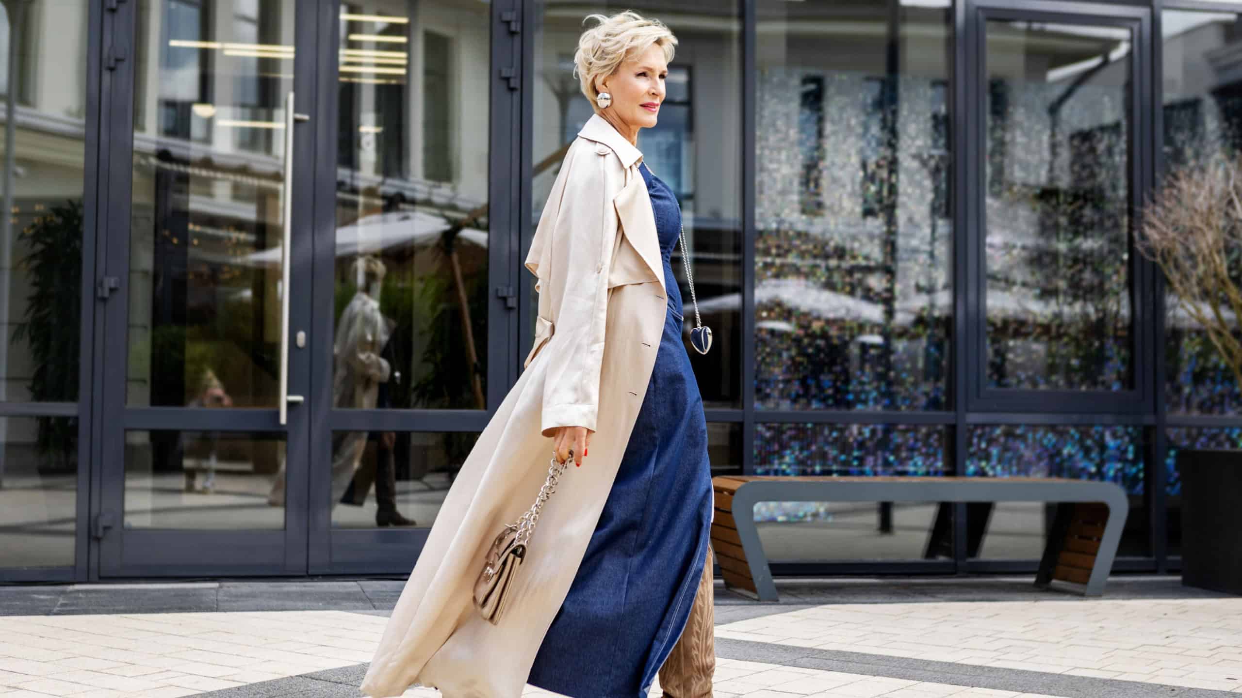 Beautiful Woman Wearing Fashionable Spring Or Fall Clothes, beige trench coat, denim dress and high boots. Female stylish Model walking Street. Autumn trend street fashion style. Outdoors