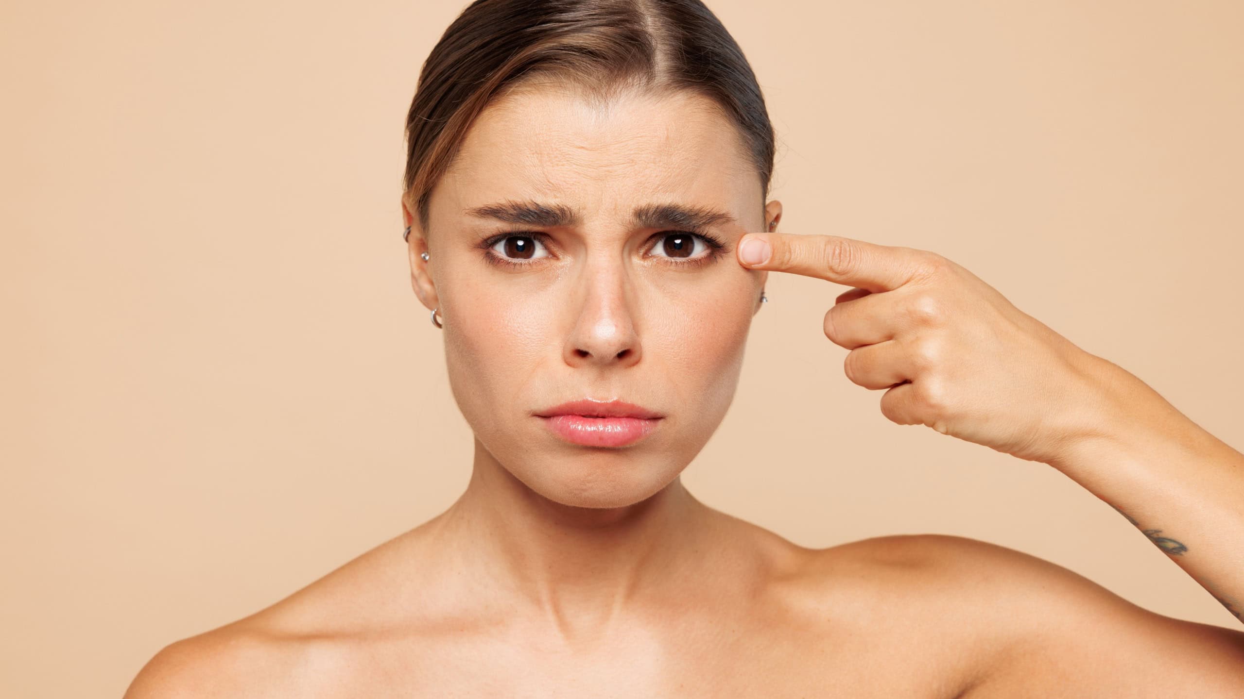 Close up sad beautiful half naked topless young woman 20s with nude make up point index finger on isolated on beige color background studio portrait. Skin care healthcare cosmetic procedures concept