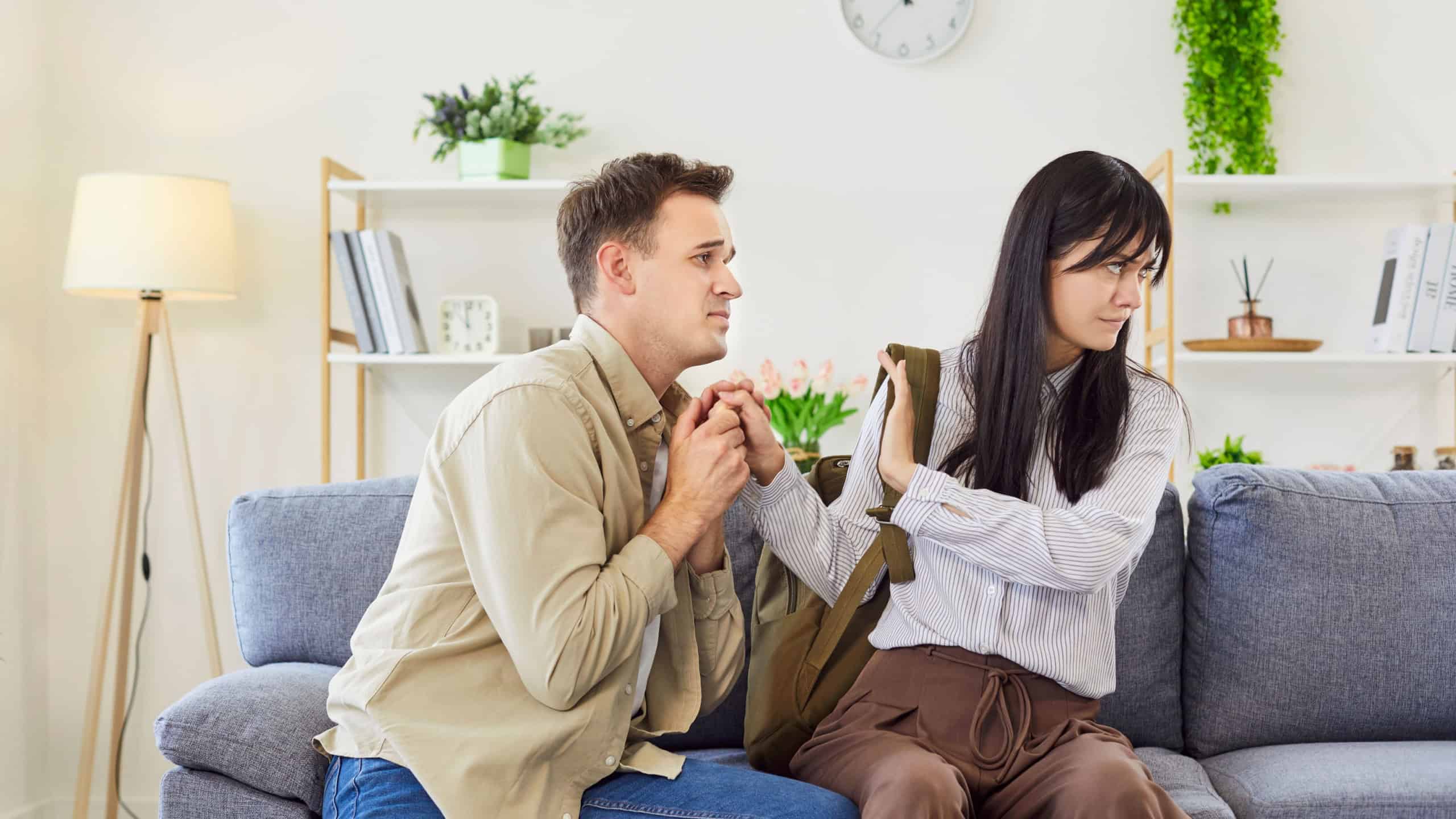 A desperate man sits beside a woman with packed bag in a living room and begging her to stay. The emotional scene highlights a moment of desperation and painful breakup after quarrel.