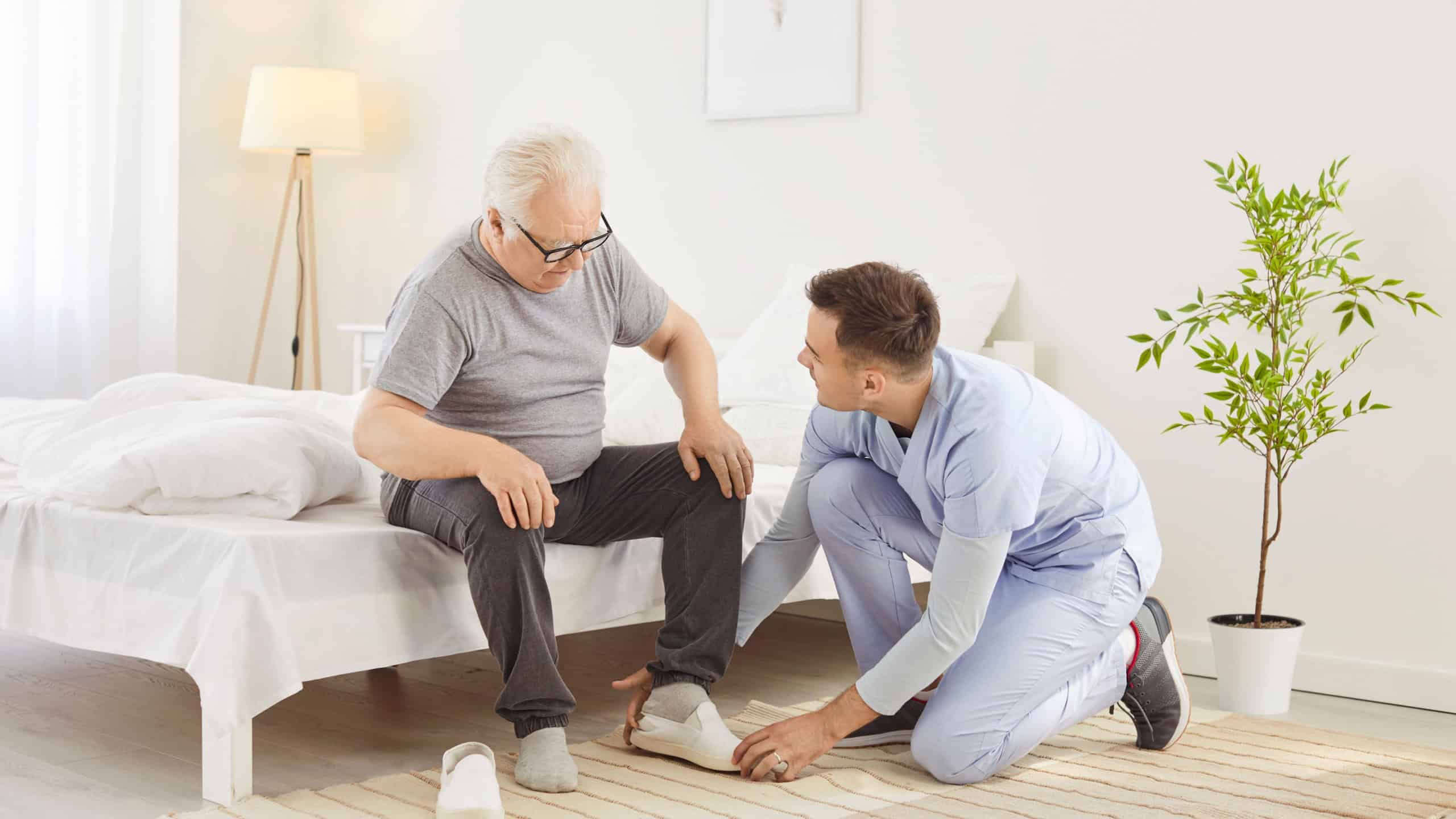 Friendly male nurse helping senior man sitting on bed to put on his shoes. Young male caregiver assisting elderly male patient with mobility and flexibility problems during everyday routine in clinic.