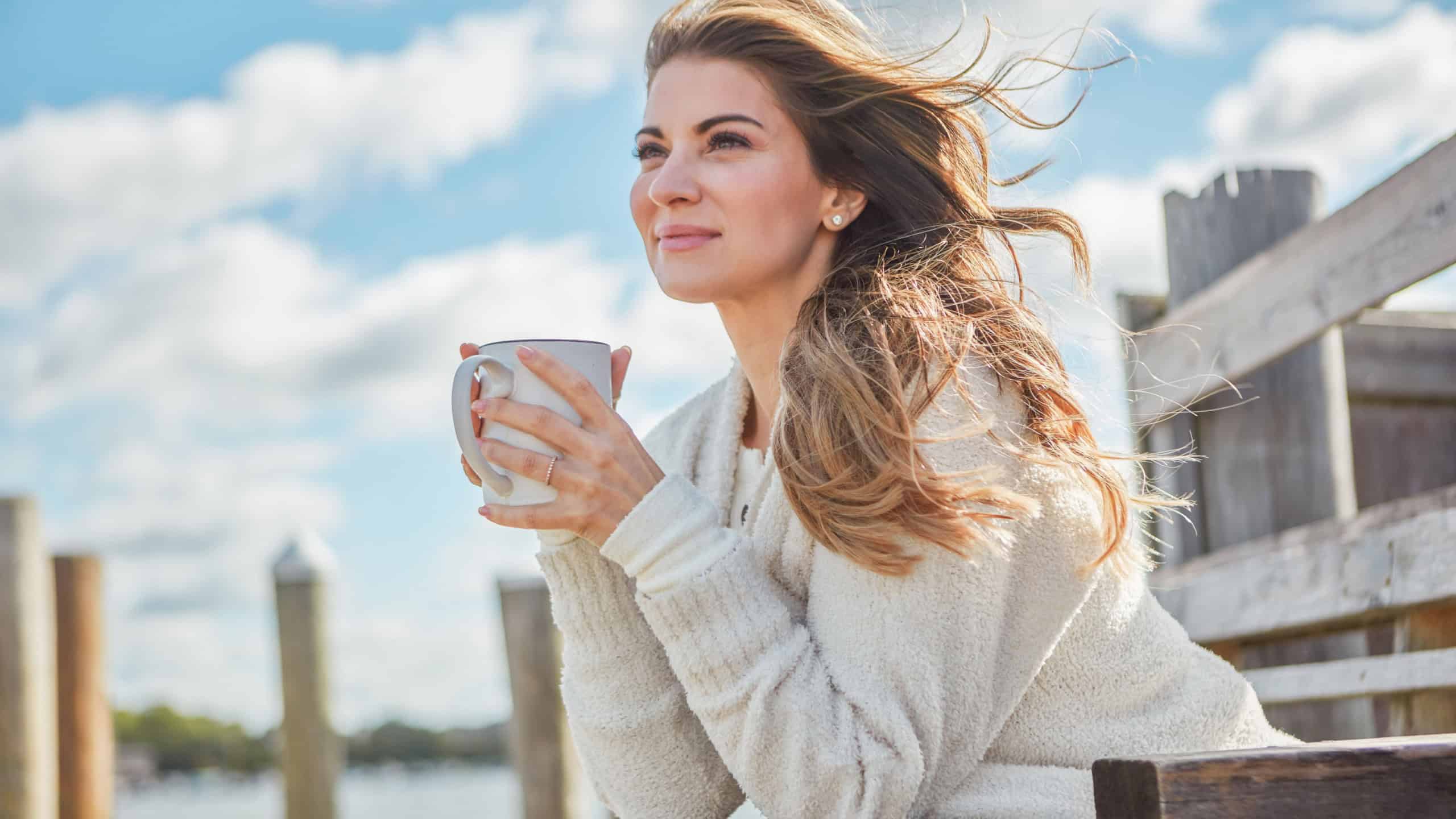 Thinking, bench and woman with coffee cup on pier, holiday reflection and positive mindset for peace. Calm, perspective and person with smile for vacation, tea drink and outdoor for daydreaming