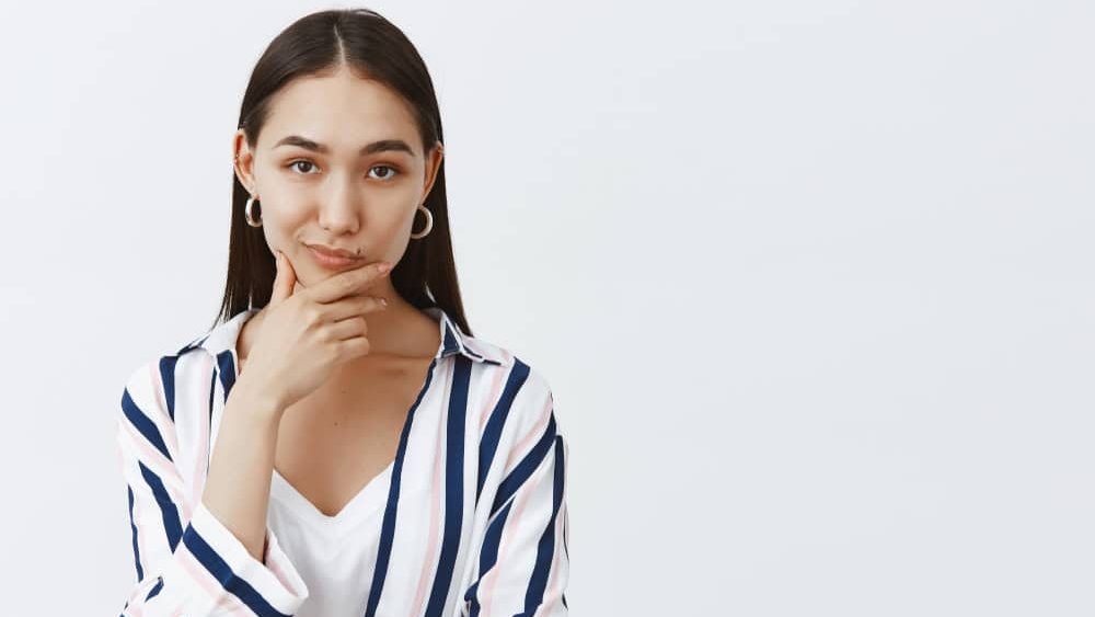 Hmm, let me think. Portrait of thoughtful smart and creative fashionable female with long dark hair, rubbing chin and smirking at camera while making up plan or choice, deciding something