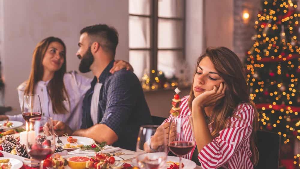Lonely woman spending Christmas Eve with friends at home, sad while sitting over Christmas dinner surrounded with couples in love