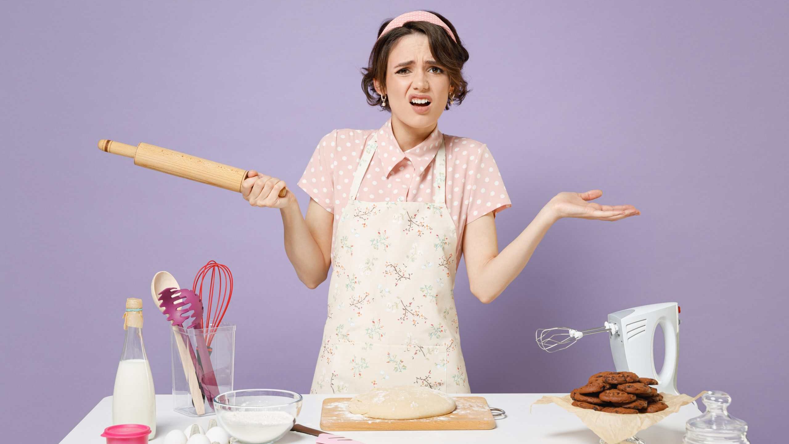 Young doubtful confused housewife housekeeper cook chef baker woman in apron work at table kitchenware hold wooden rolling pin spread hands isolated on violet background Process cooking food concept