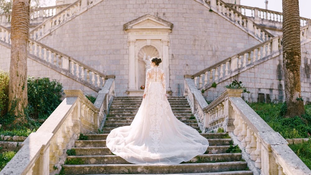 Bride in white full-length dress stands on stone steps in a green garden