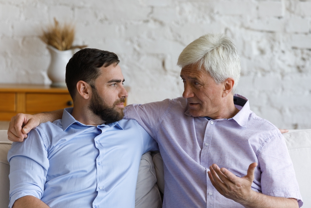 Elder grey haired loving father giving support and advice to grownup son. Senior parent and adult child talking on couch at home, discussing family life, keeping trusting relationships