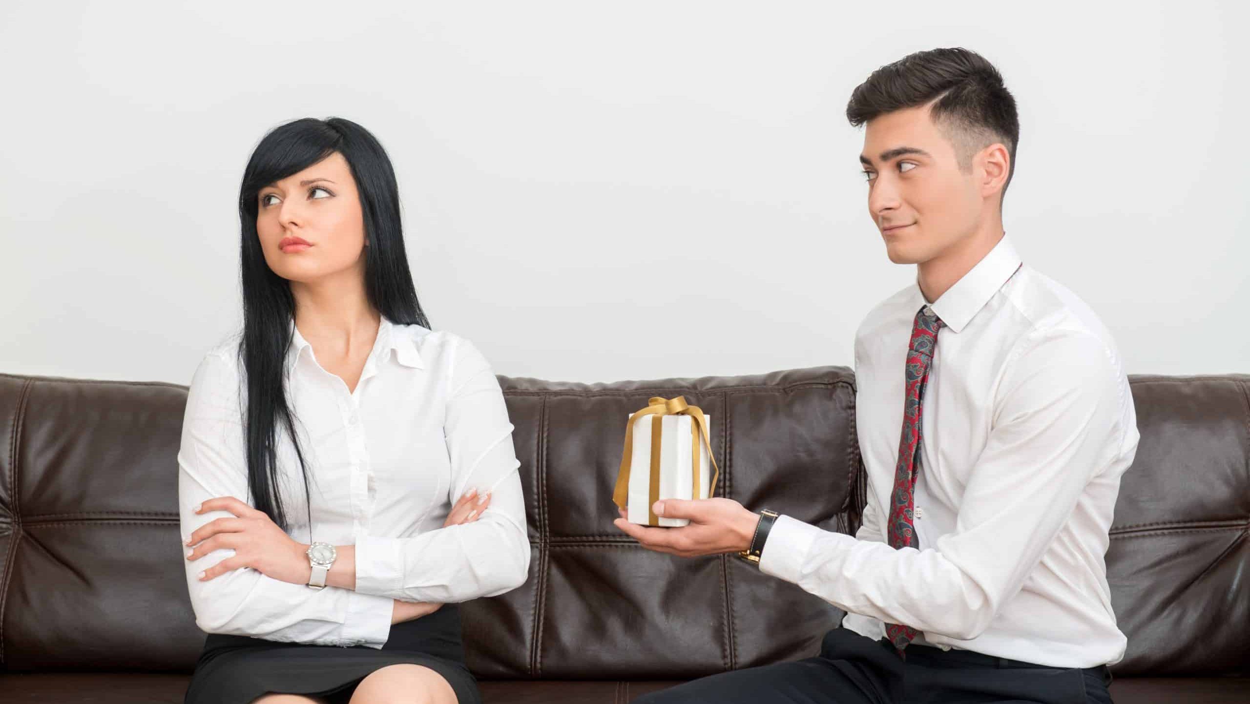 Side view of Business couple in formal clothing sitting on sofa in office, man flirting and giving white gift Box to dissatisfied and unhappy woman that looks aside