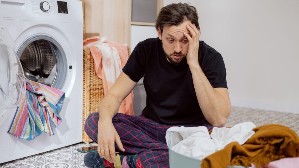 Guy sits on the laundry room floor with the washing machine open, throwing all the clothes into the drum, guy doesn't know how to do housework, face crumpled, catches head with hand.