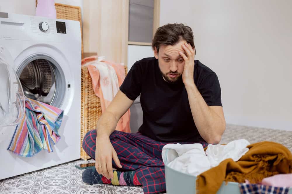 Guy sits on the laundry room floor with the washing machine open, throwing all the clothes into the drum, guy doesn’t know how to do housework, face crumpled, catches head with hand.