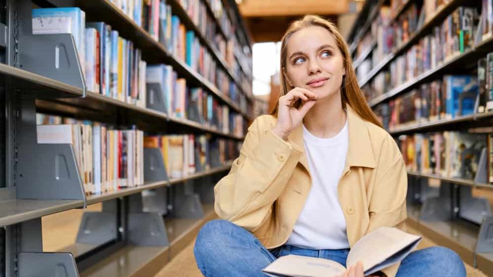 Smart pretty creative girl student holding book sitting on floor among bookshelves in modern university campus library looking away thinking of college course study thinking reading literature.