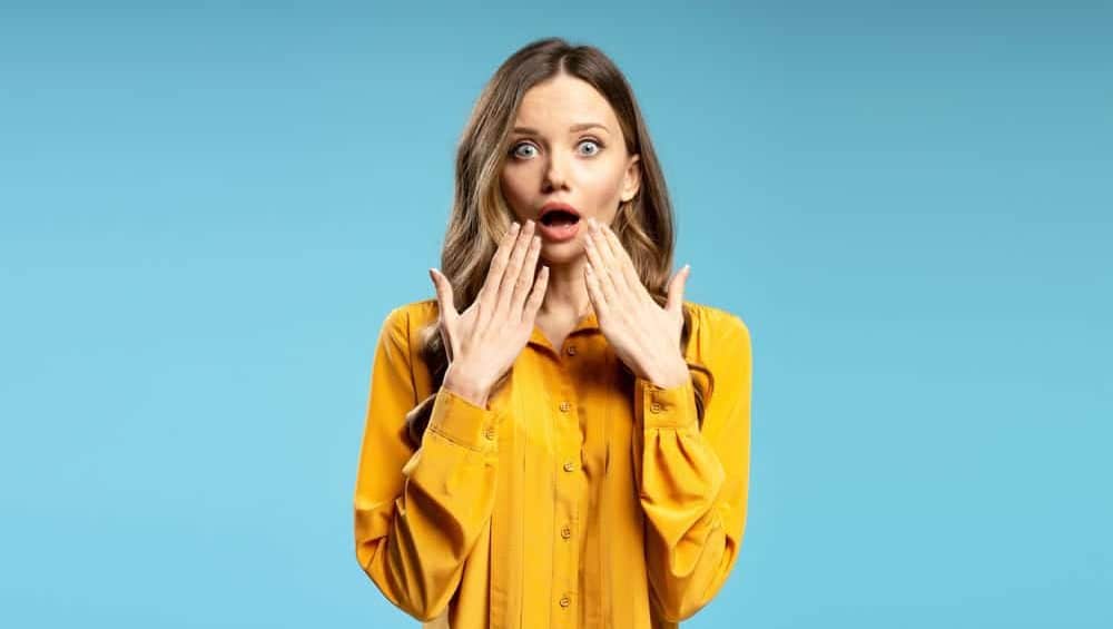 Studio portrait of surprised businesswoman covering her mouth with hands, expressing astonishment and disbelief on blue background