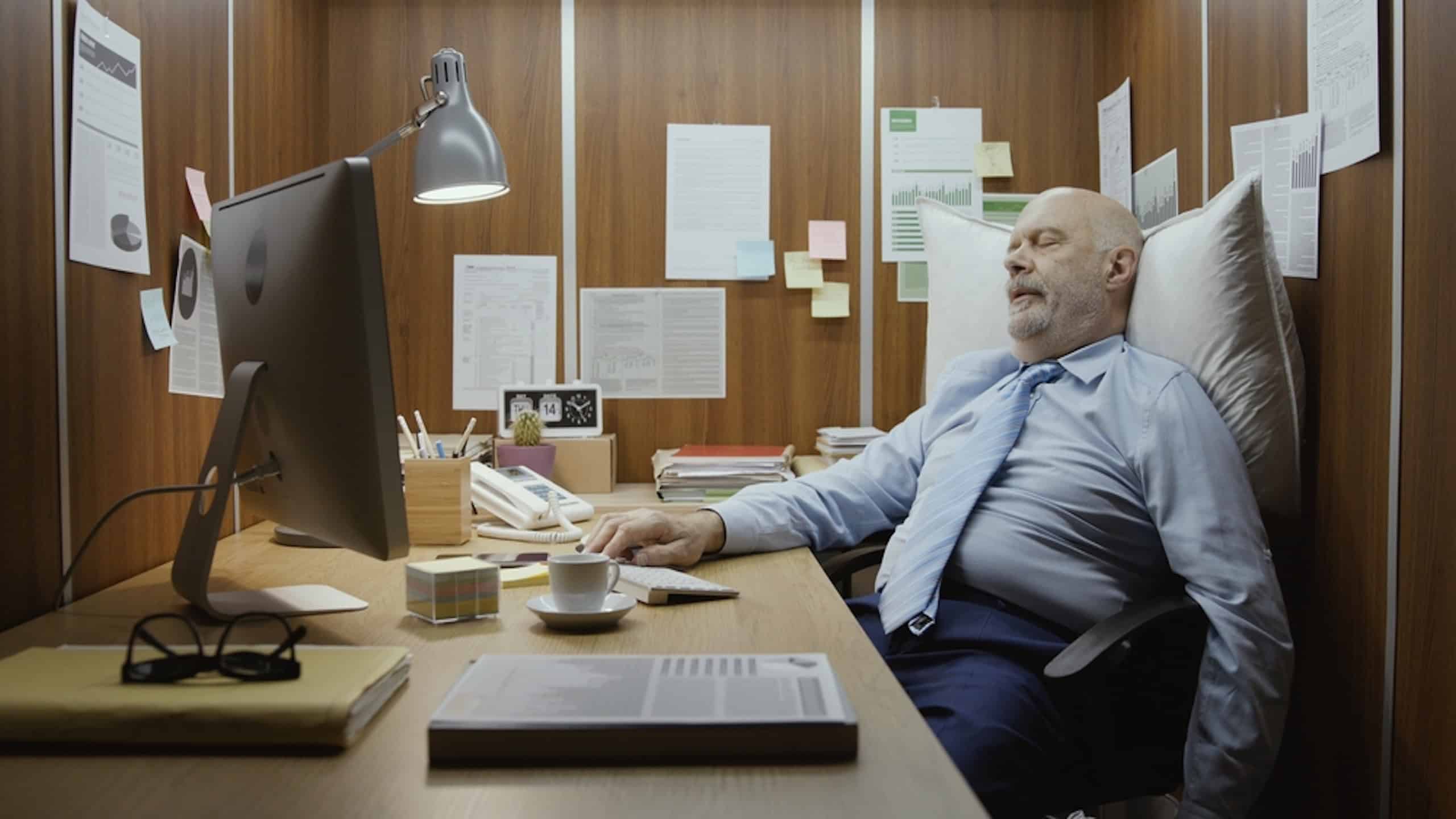 Lazy senior office worker sleeping at his desk in a tiny office, he is leaning on a pillow and resting