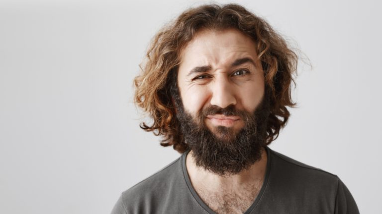 Man is not sure, having doubt. Portrait of attractive curly-haired husband frowning and grimacing from dislike and displeasure, hearing something ridiculous or dumb while standing over gray wall