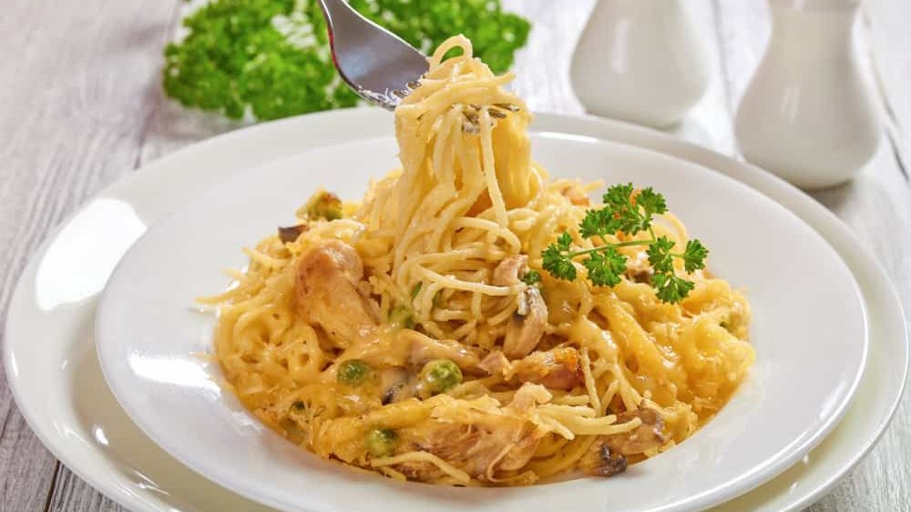 female hand is holding a fork with portion of Tetrazzini, classic american casserole of paste, poultry meat mushrooms and cheese, view from above, close-up