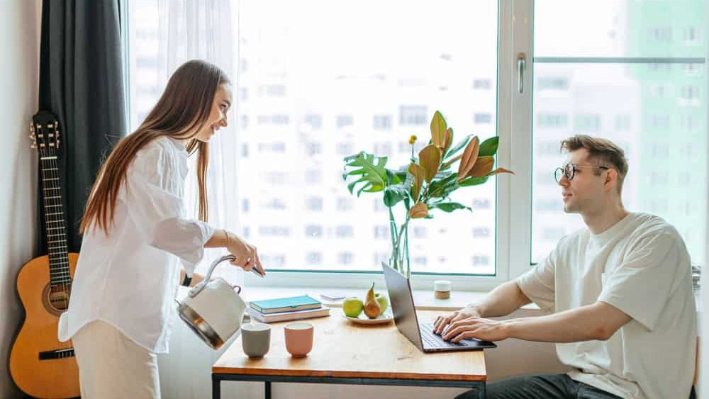 beautiful young caucasian woman pouring tea to working husband, man work from home using laptop. merciful kind woman support him, want to give him a cup of tea
