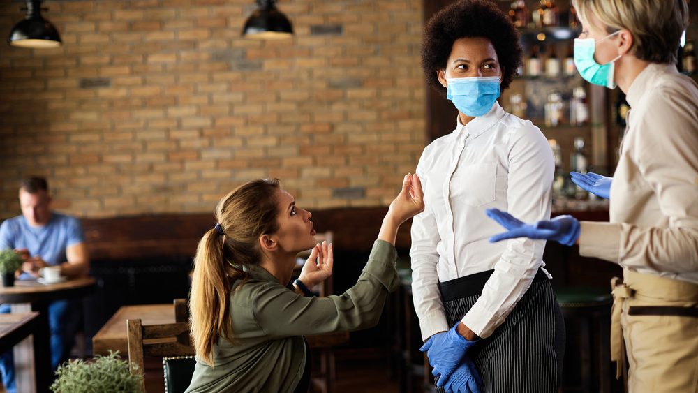 Upset woman having an argument with waitresses because she doesn't want to wear protective face mask in a cafe.
