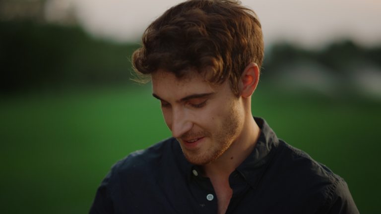 Portrait of thoughtful man feeling shy outdoors. Closeup thinking guy posing on meadow. Young male person looking at camera at summer park. Handsome man standing outside at sunset.