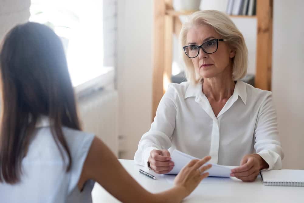 Pensive middle-aged businesswoman listen to young employee or job candidate at interview in office. Confident senior female boss or recruiter talk speak with work applicant. Employment concept.