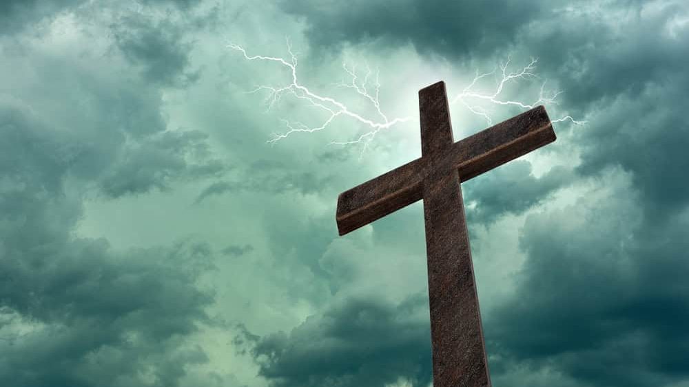 Dramatic image showing a religious church cross framed against a sky of lightning, bright light and sun rays, showing the coming of the Lord. Copy space available on the left of the cross.
