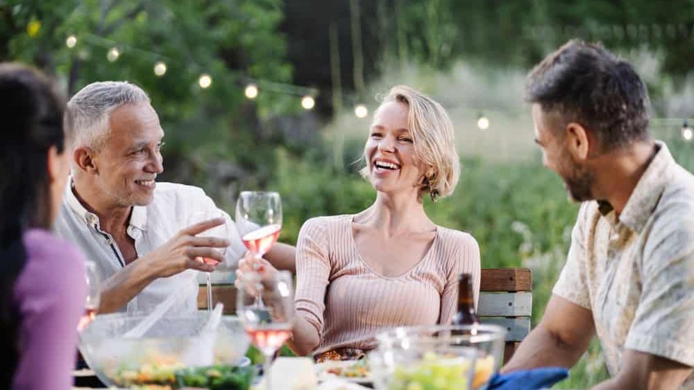 Mature friends having dinner around table in a garden on a summer evening, drinking wine, having fun talking and laughing