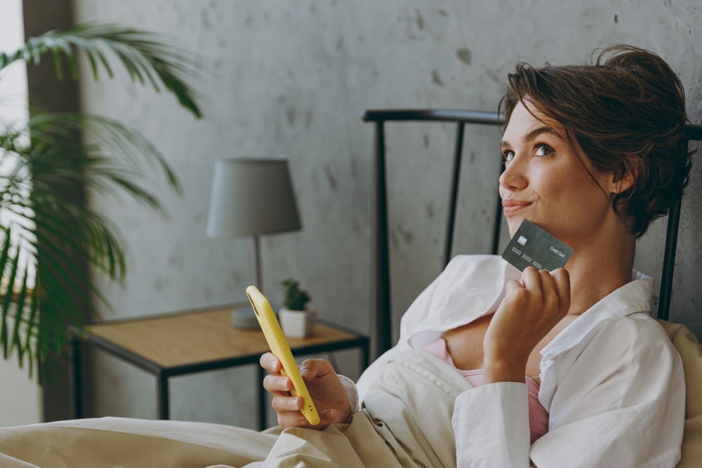 Side view young woman wear white shirt pajama she lying in bed use mobile cell phone credit bank card shopping online order delivery rest relax spend time in bedroom lounge home in hotel room wake up