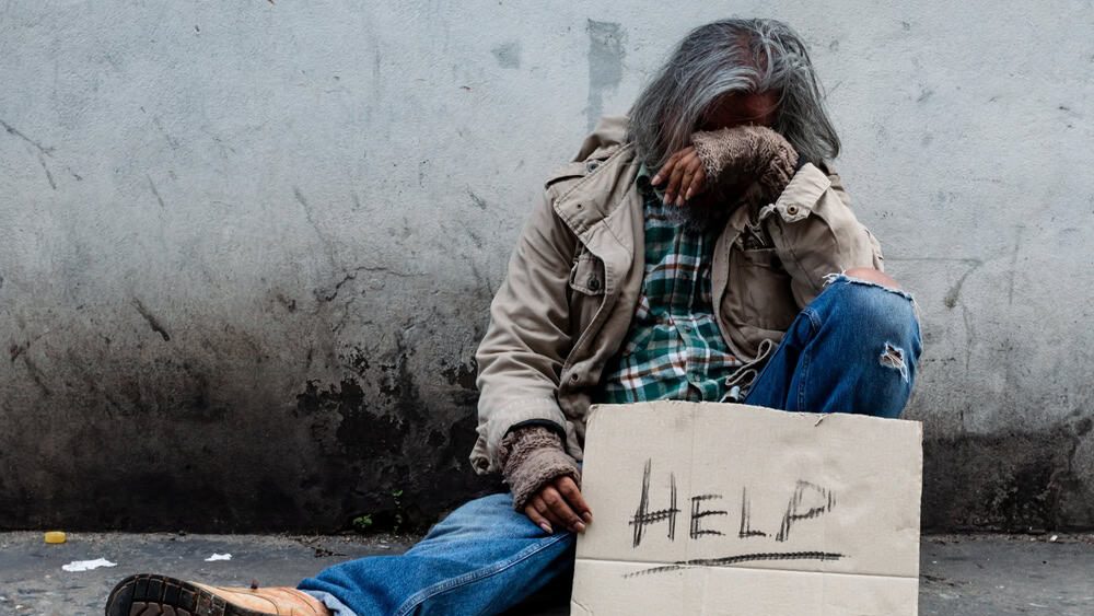 homeless, long-haired Asian man sits hopelessly leaning against a wall as there is no one to help him with work and food in his hand holding a sign for help. Homeless sleep on streets