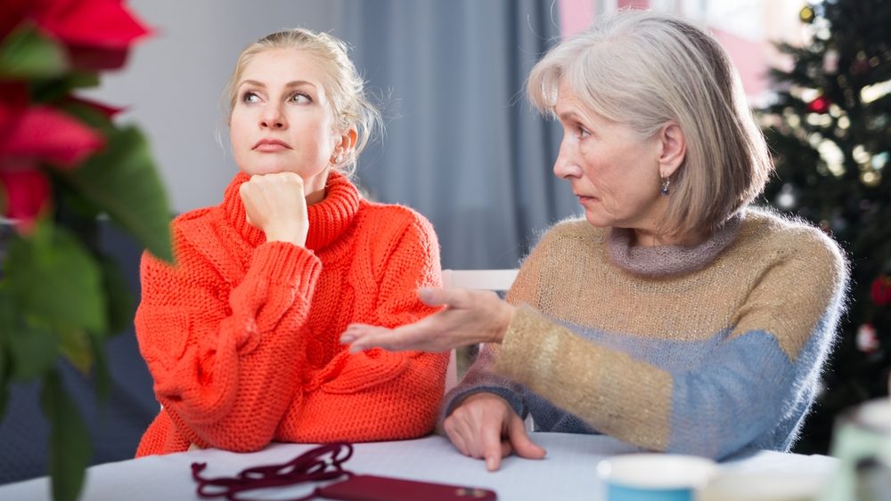 Mature mother explains something to her adult thoughtful daughter, while sitting at the table in the room